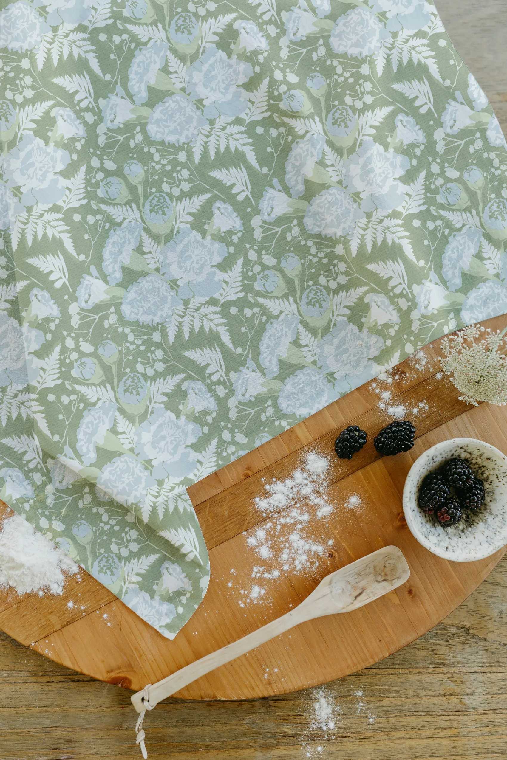 A wooden cutting board with a small bowl of blackberries, a wooden spatula, and scattered flour, with a floral-patterned cloth partially covering the board.