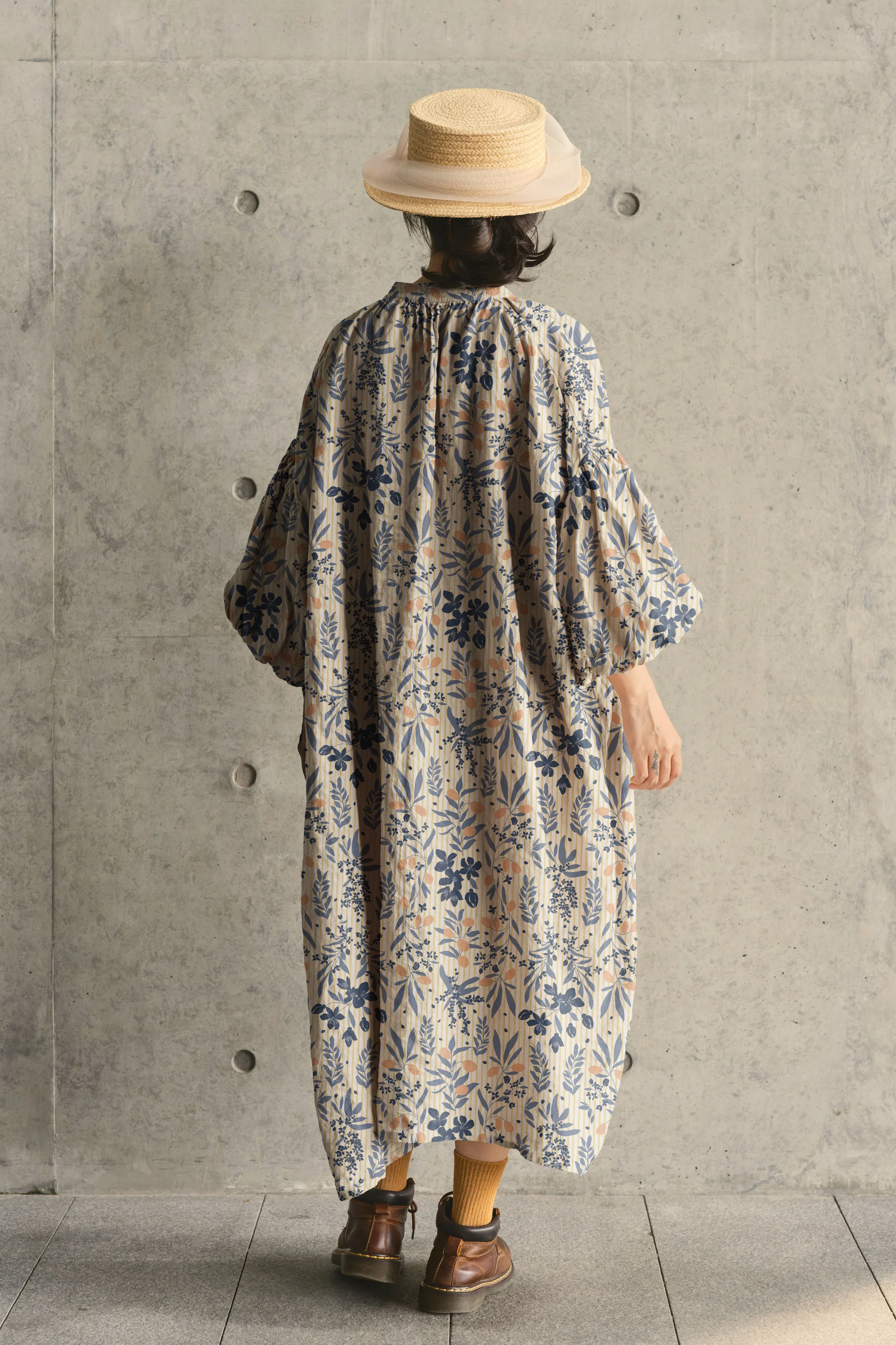 A person standing with their back to the camera, wearing a large floral patterned dress, brown boots, yellow socks, a wide-brimmed straw hat with a white ribbon, and standing against a concrete wall.