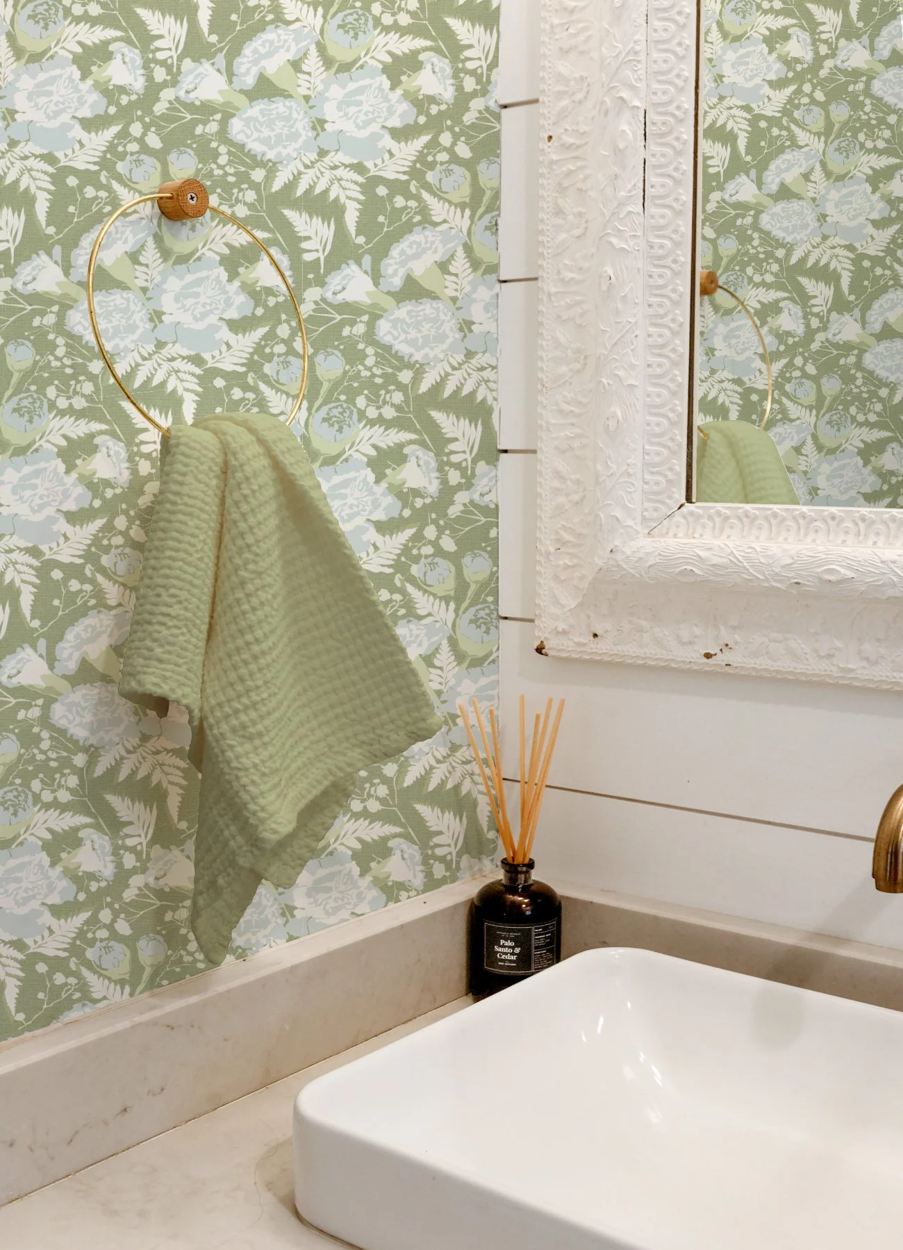 A bathroom with a floral patterned wallpaper, a mirror with a decorative white frame, a yellow textured towel hanging on a gold ring, a black jar with wooden sticks on a white sink countertop, and a reed diffuser.