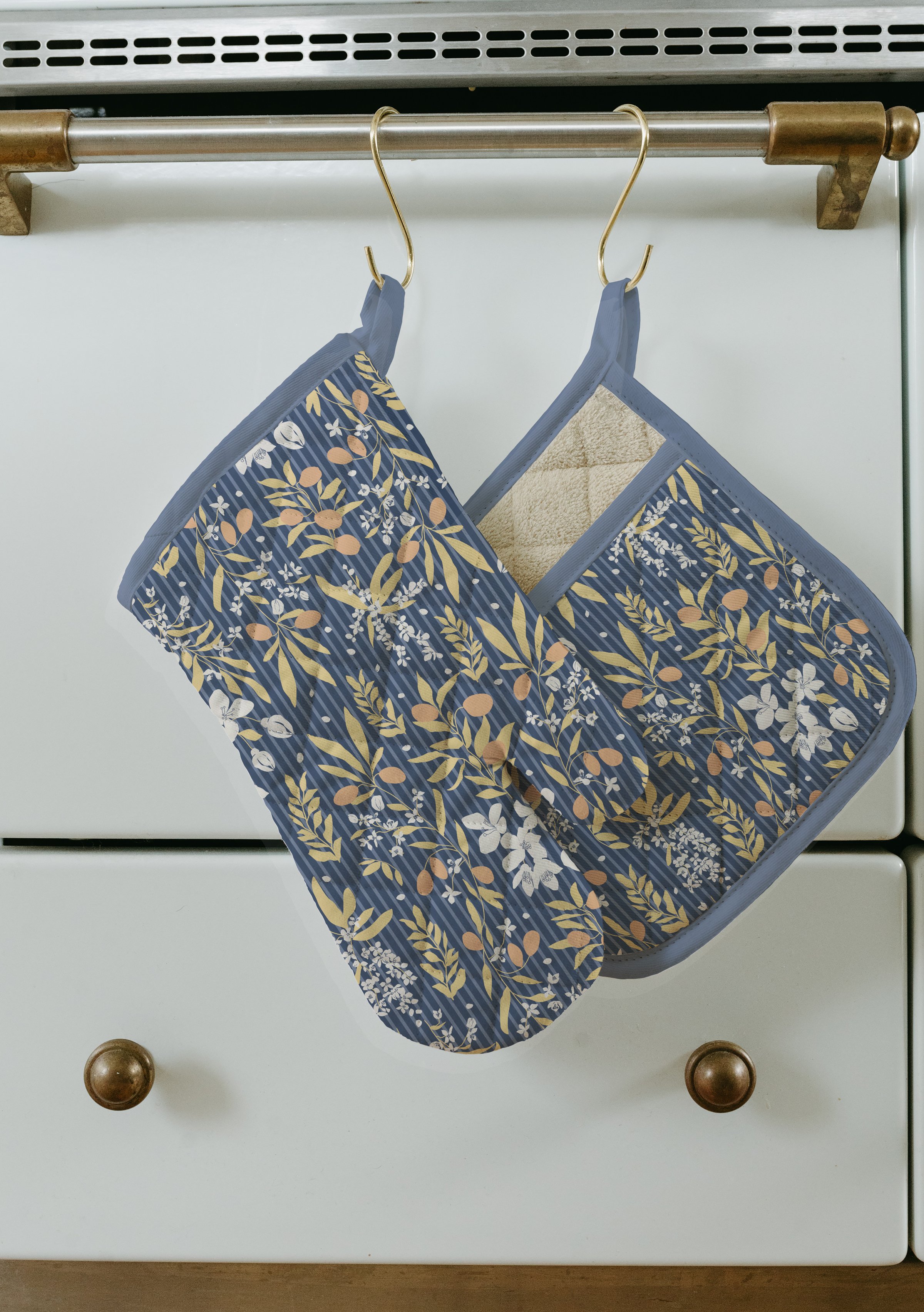 Two patterned oven mitts hanging on a metal rod on a white piece of furniture with wooden knobs.