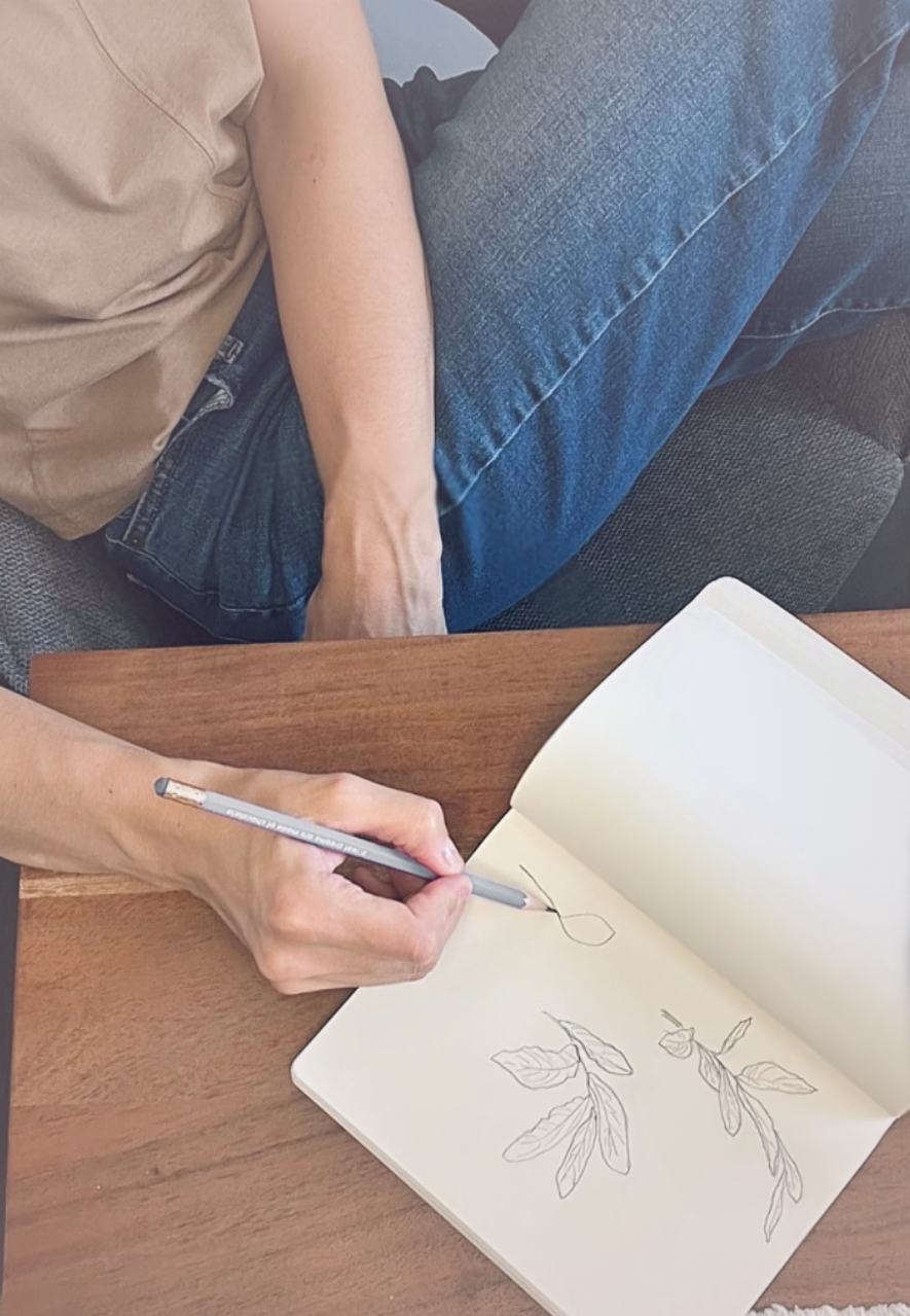 A person sitting at a wooden table, drawing botanical sketches of leaves and branches in a sketchbook with a pencil.