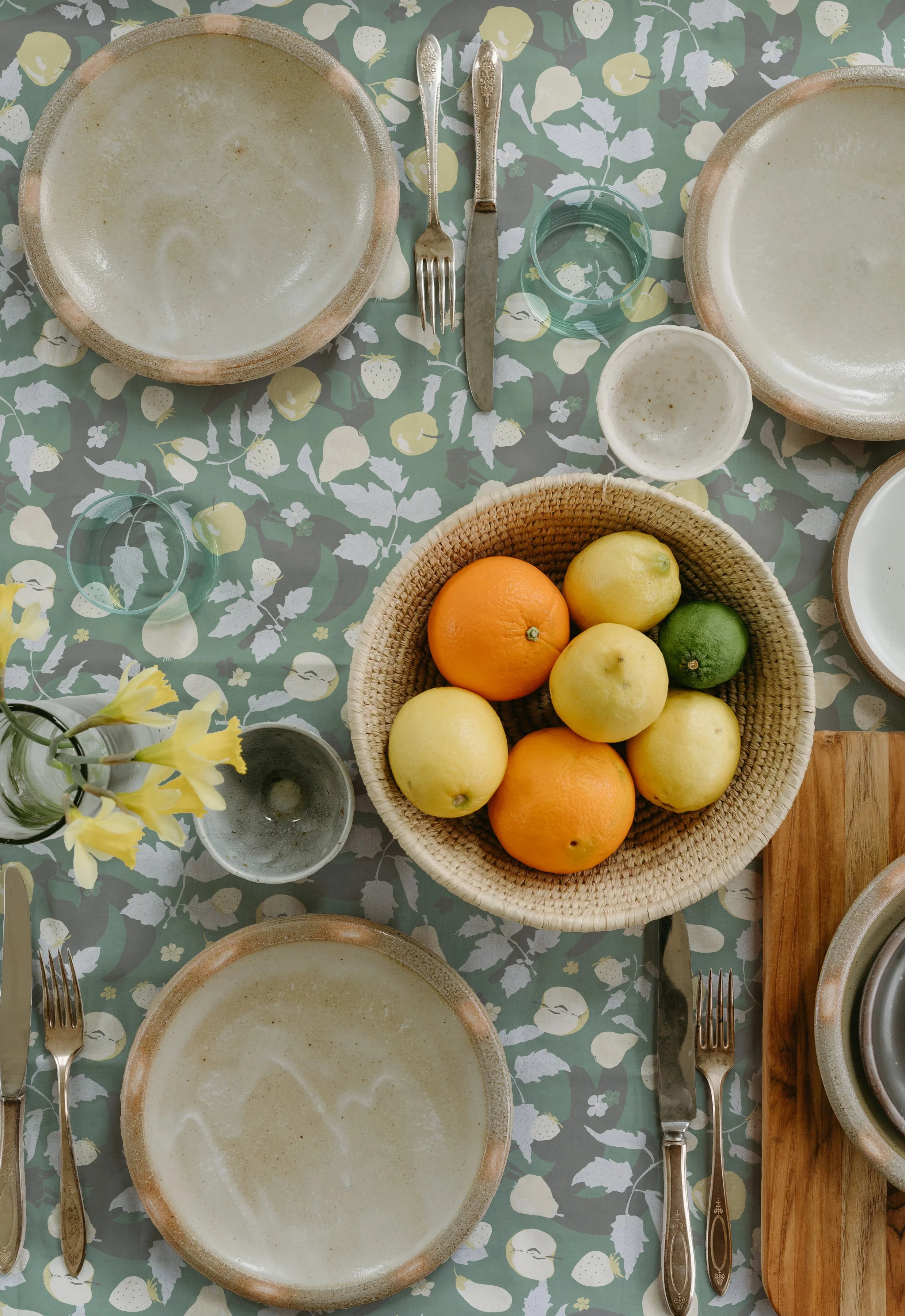 A table set with empty ceramic plates, forks, knives, glasses with drink, and a basket of lemons, oranges, and a lime. The tablecloth has a floral fruit pattern.