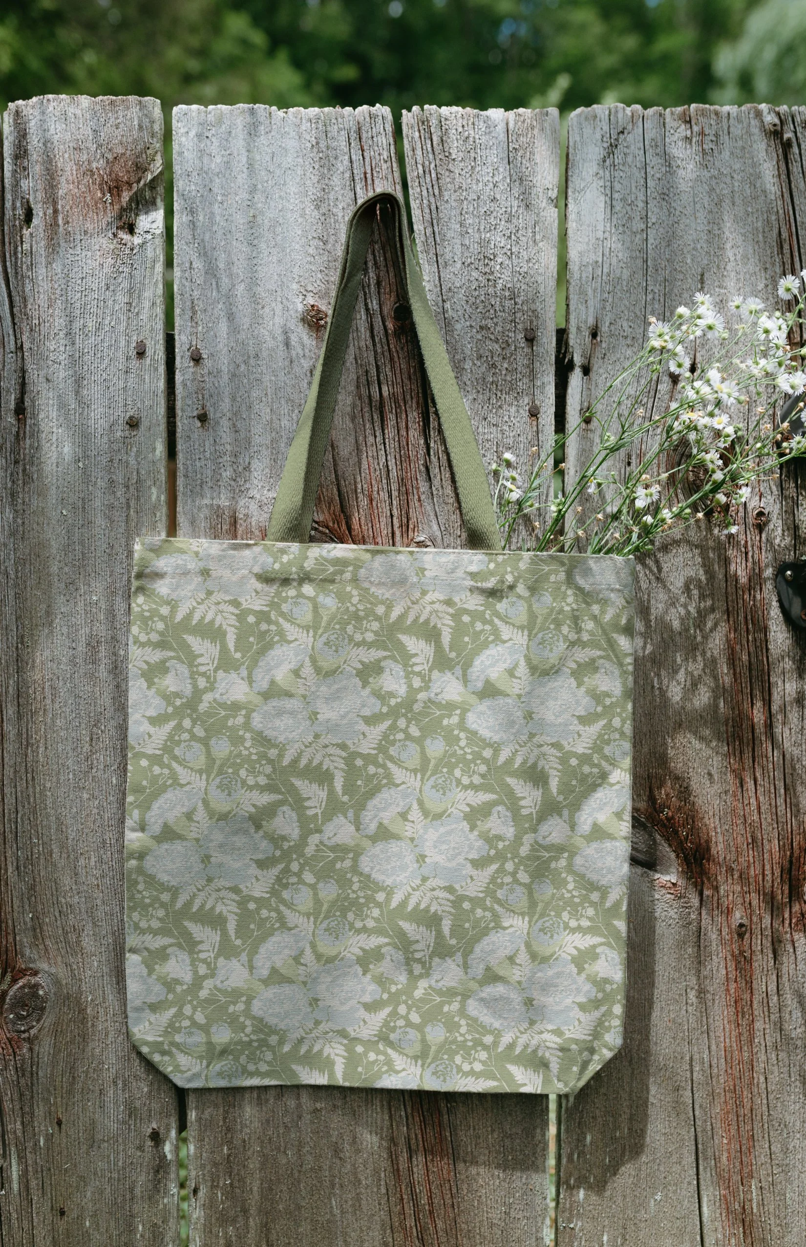A green reusable tote bag with a white floral pattern hanging on a weathered wooden fence, with some white daisies below the bag.