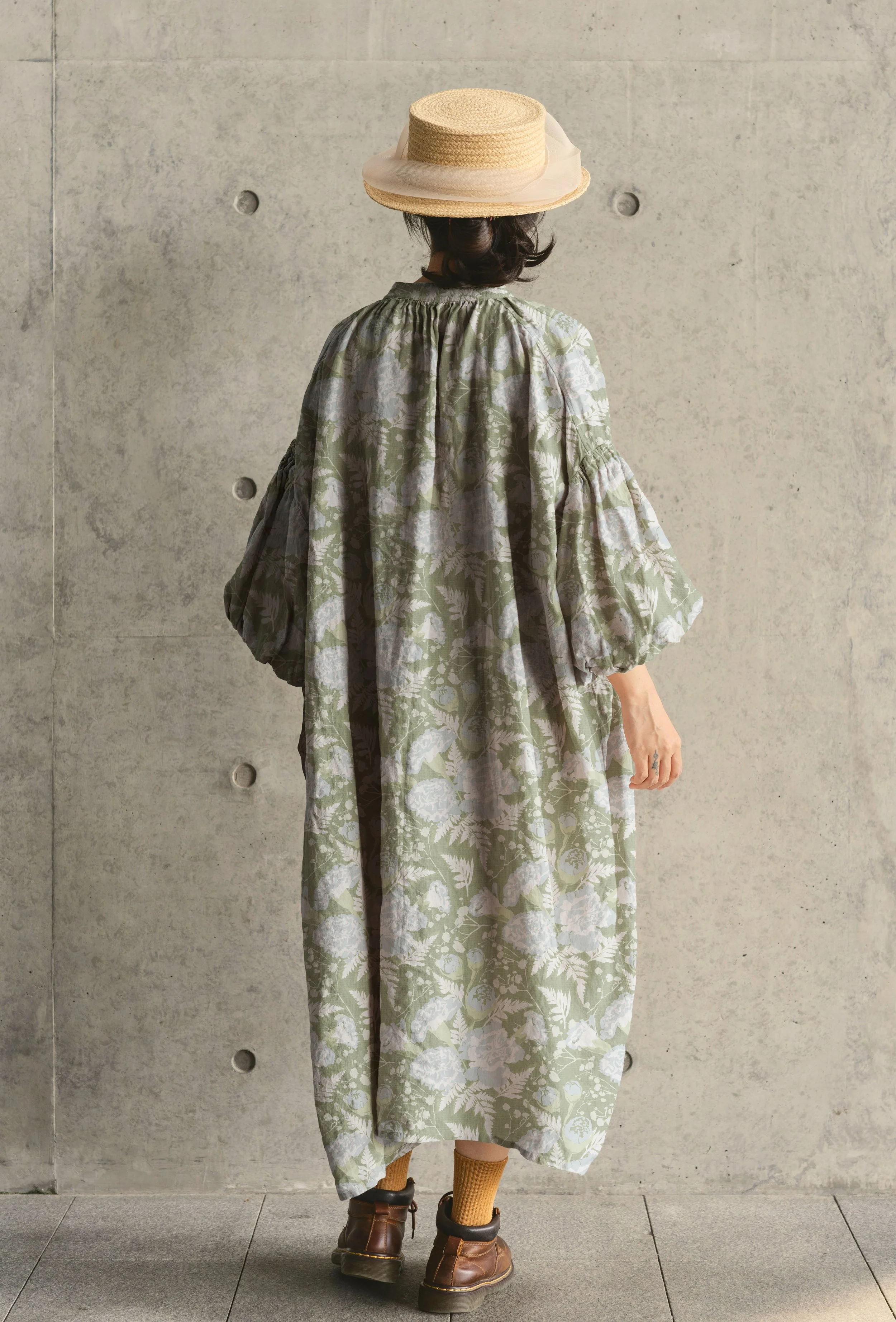 Person wearing a wide-brimmed straw hat, a floral-patterned dress, brown boots, and mustard-colored socks, standing against a concrete wall, viewed from behind.