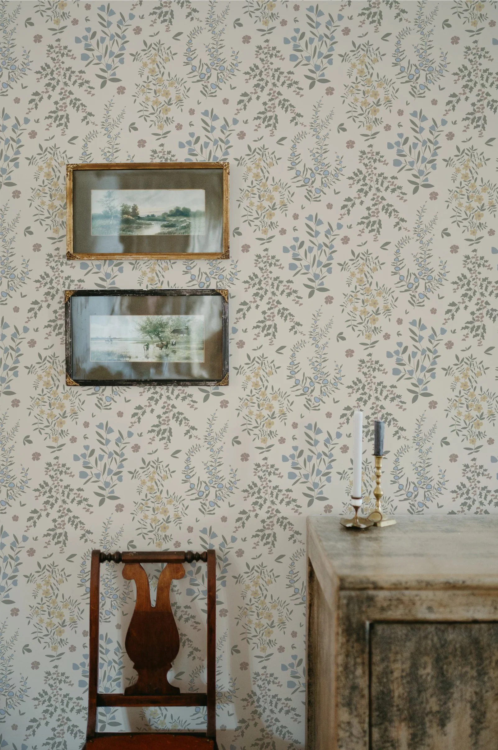 A vintage-style room with wallpaper featuring a floral and leaf pattern in muted colors. On the wall, there are two framed landscape paintings, one above the other. In front of the wall is a wooden chair with a carved backrest and a top rail. To the right, a distressed wooden table holds two candlesticks with candles.