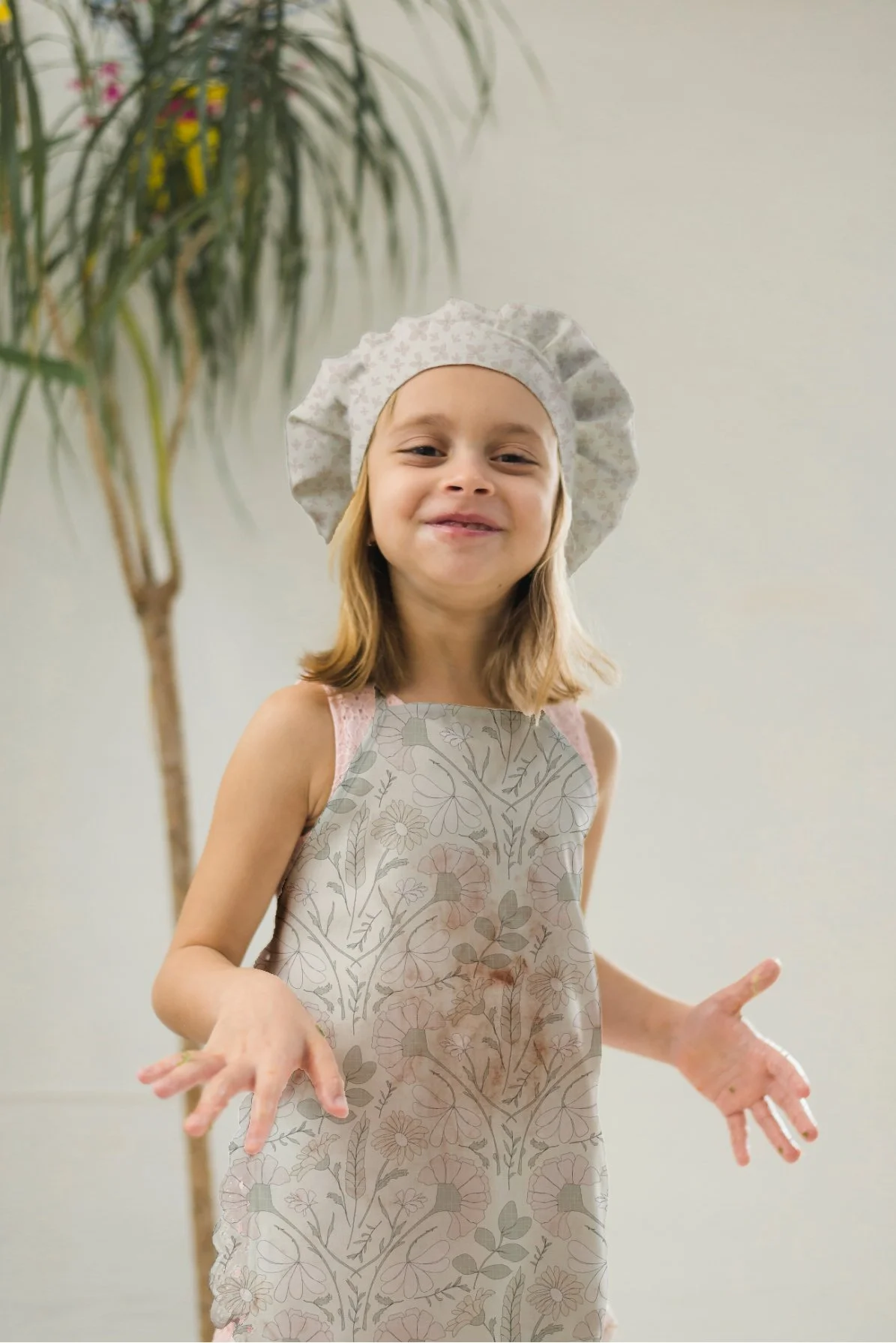 A young girl wearing a floral apron and a patterned chef's hat, standing indoors near a potted plant, smiling with her hands slightly raised.