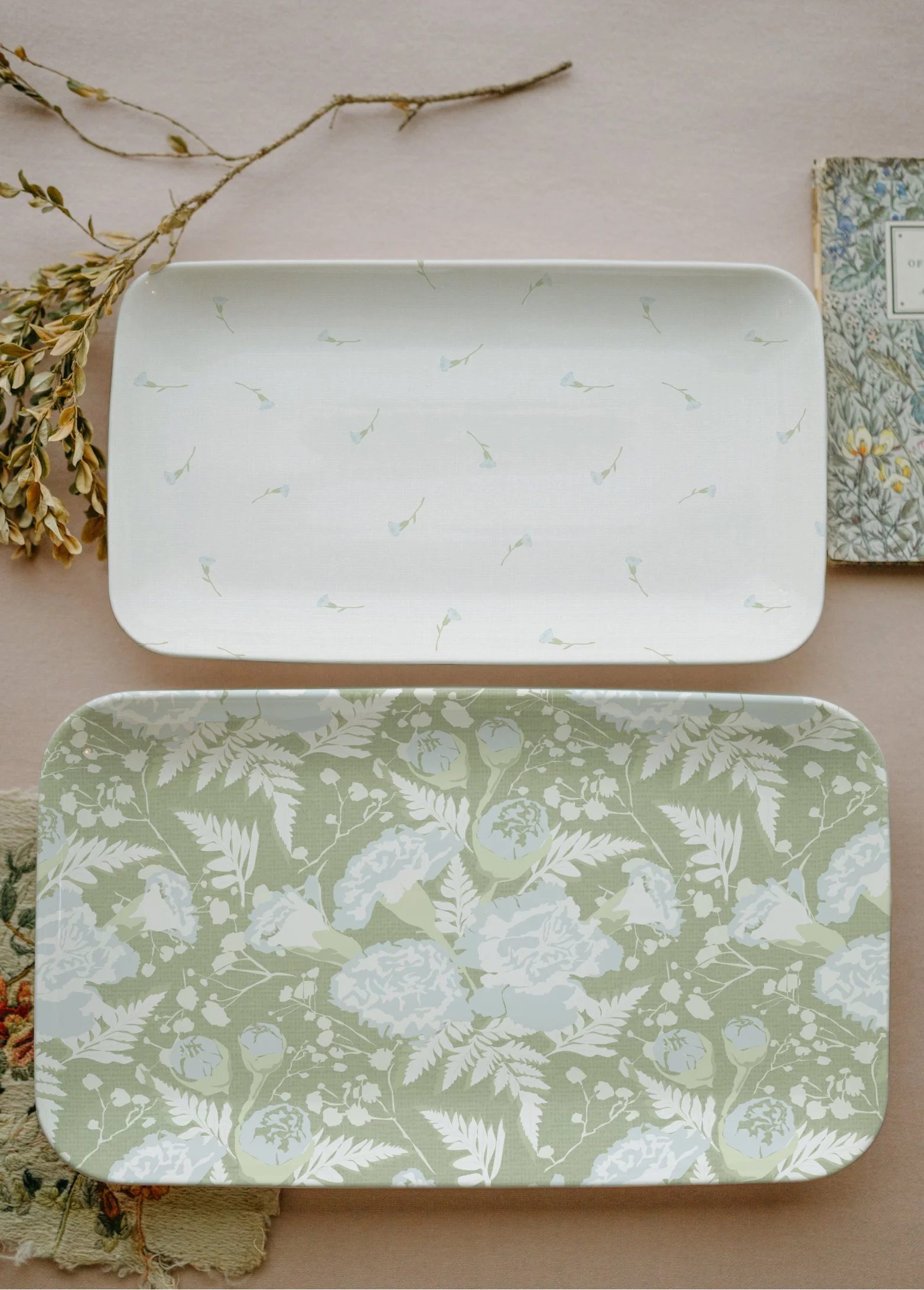 Two rectangular trays with floral and botanical patterns, one white with small white flowers and green stems, the other pale green with white leaves and flowers. Dried flowers and a book are partially visible in the background.