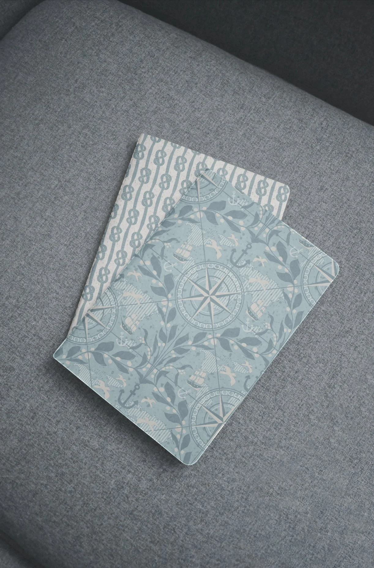 A square piece of decorative paper with nautical designs, including an anchor, compass, and ships, placed on a gray fabric surface.