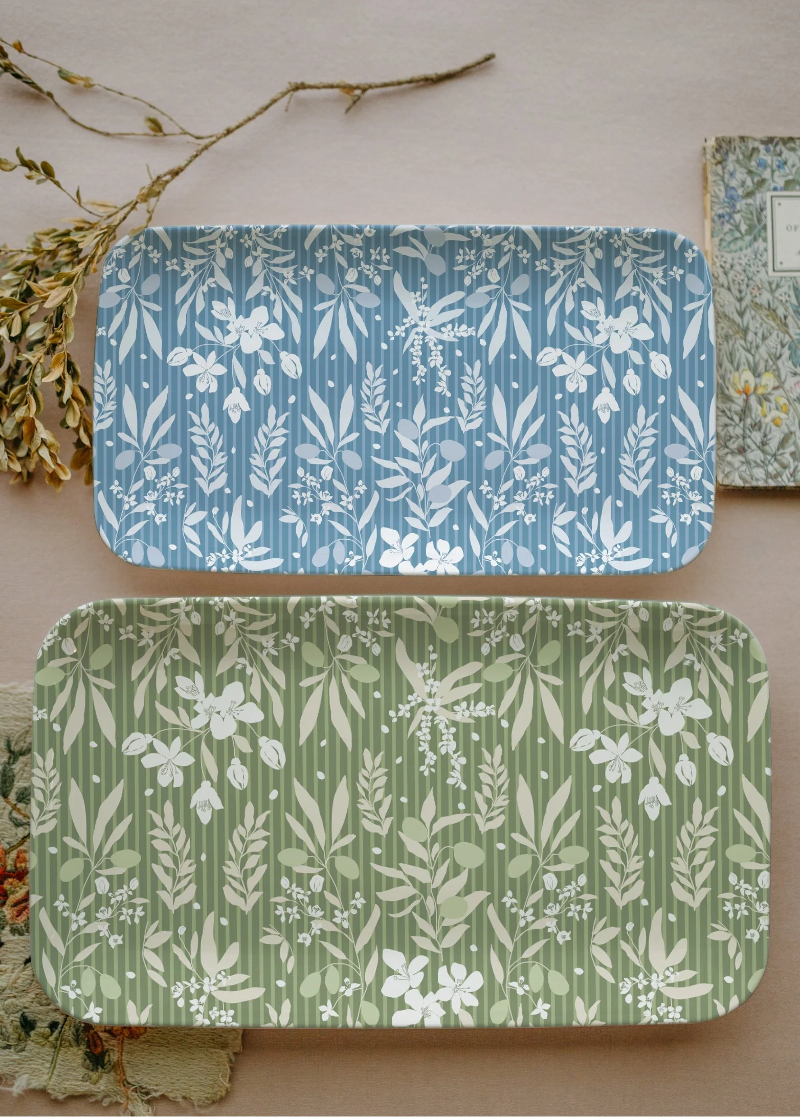 Two decorative boxes with floral patterns, one blue and one green, placed on a beige surface with dried plant stems nearby.