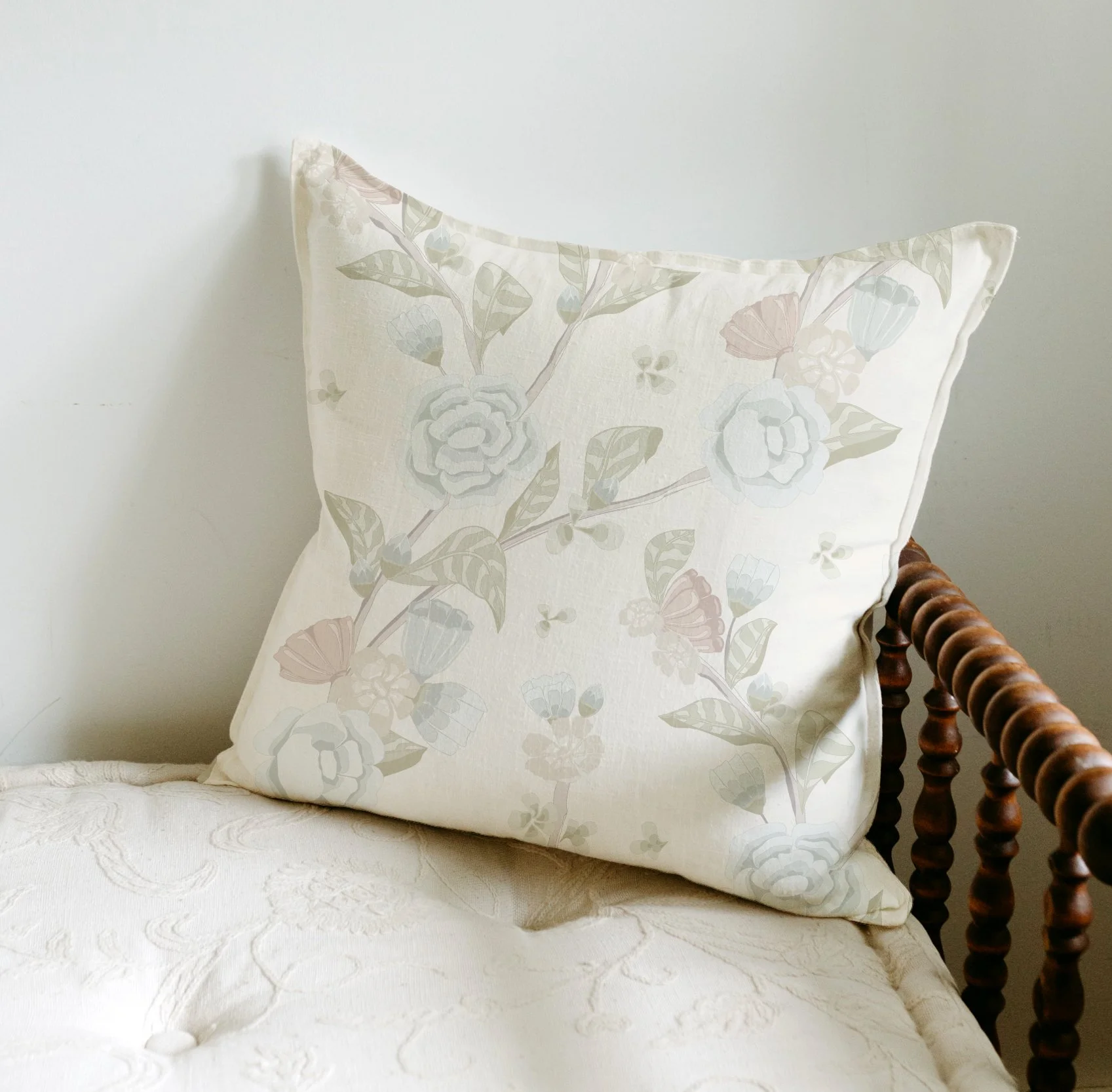 A decorative pillow with a floral pattern on a wooden chair with a white embroidered bedspread underneath.