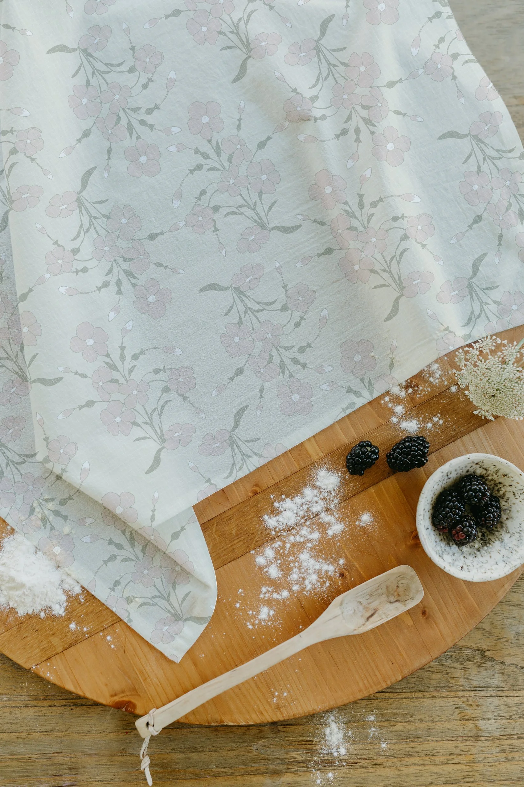 A wooden table with a sheet of paper, a bowl of blackberries, and a dish with blackberries, surrounded by scattered flour and a wooden spatula.