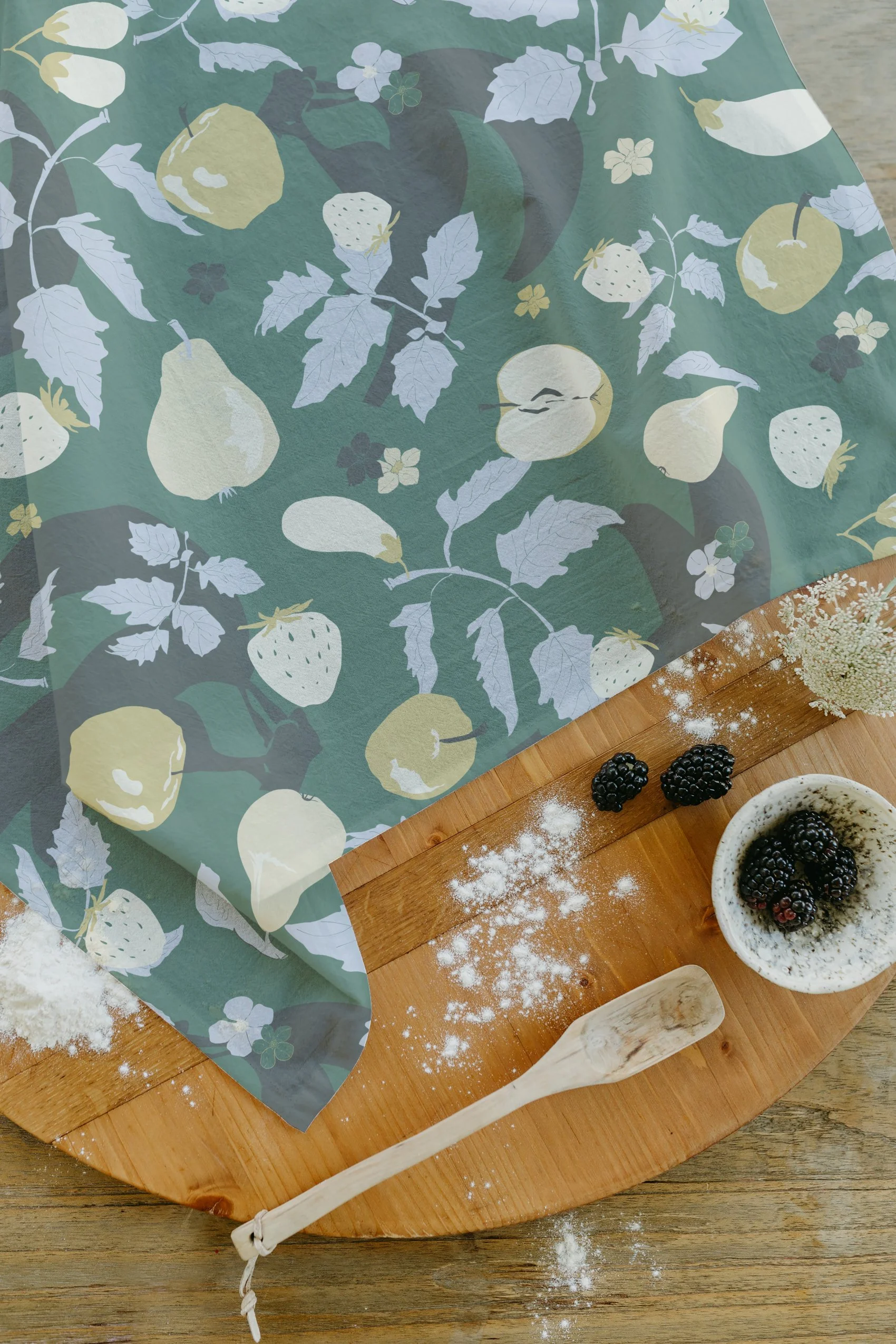 A wooden cutting board with a wooden spatula, scattered flour, a small bowl of blackberries, and a cloth with a lemon and strawberry pattern, all on a wooden surface.