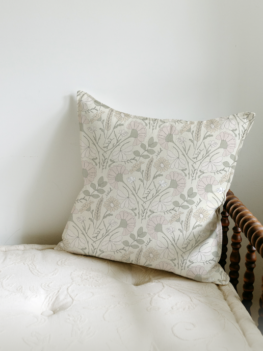 A decorative pillow with a floral pattern resting against a white wall on a bed with a cream-colored quilt. Pattern design for licensing.