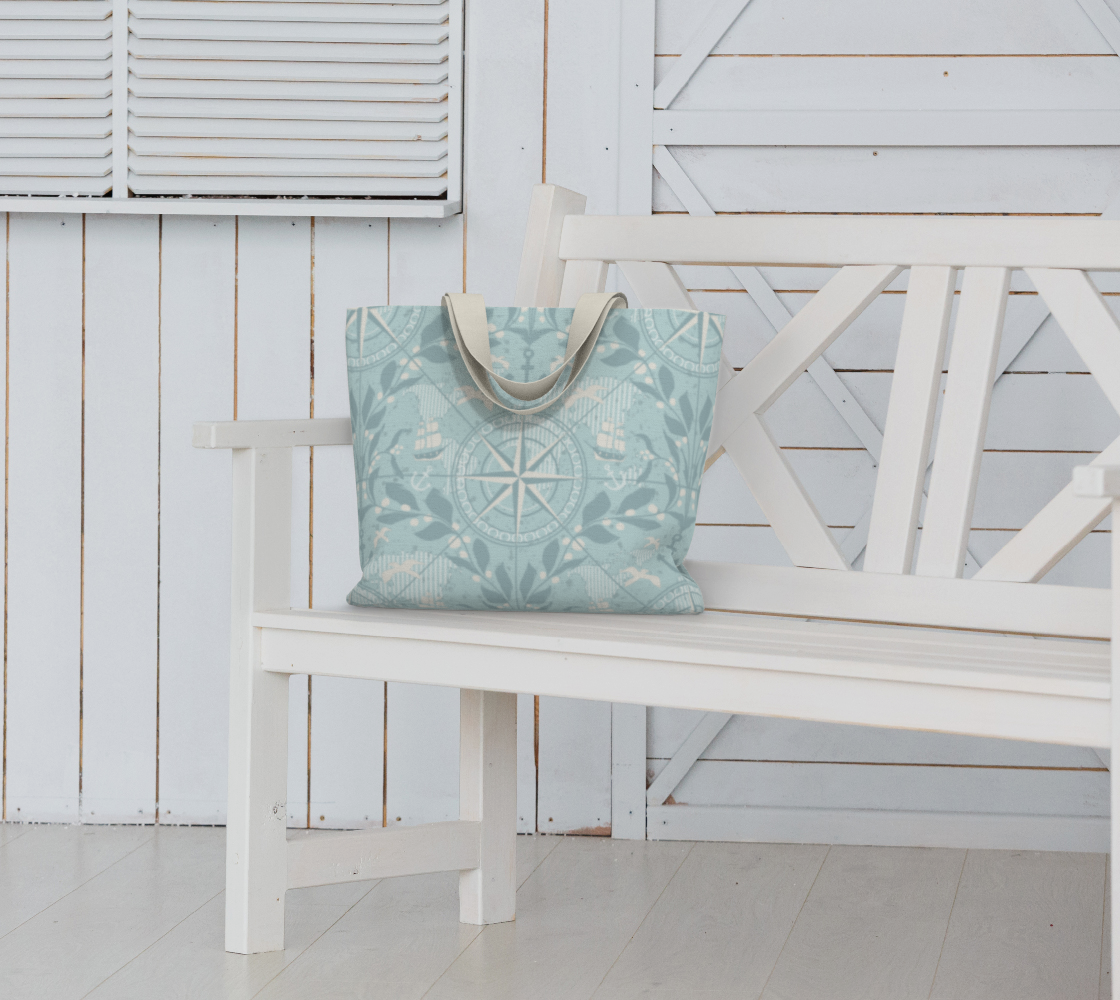 A light blue tote bag with nautical and compass designs sits on a white wooden bench in front of a white wooden wall and a window with louvered shutters.