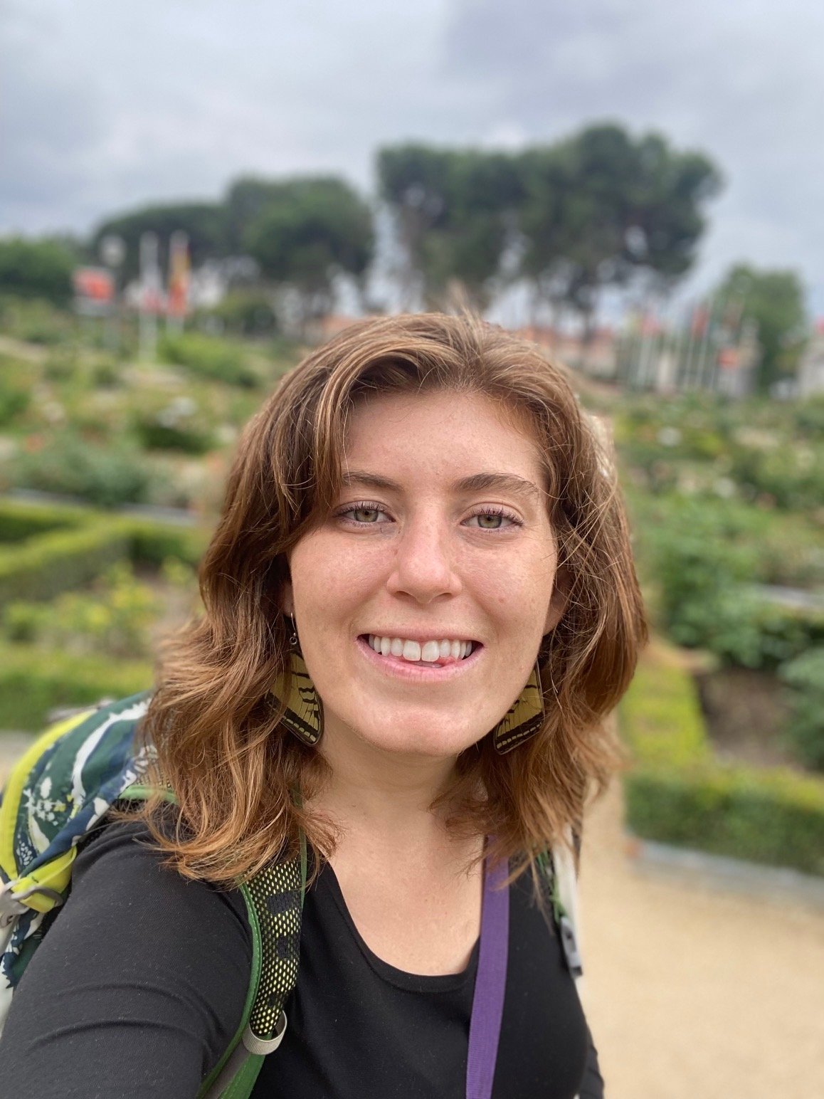 A woman with shoulder-length auburn hair smiling outdoors, wearing earrings, a black top, and a backpack, in a garden or park with green plants and trees in the background.