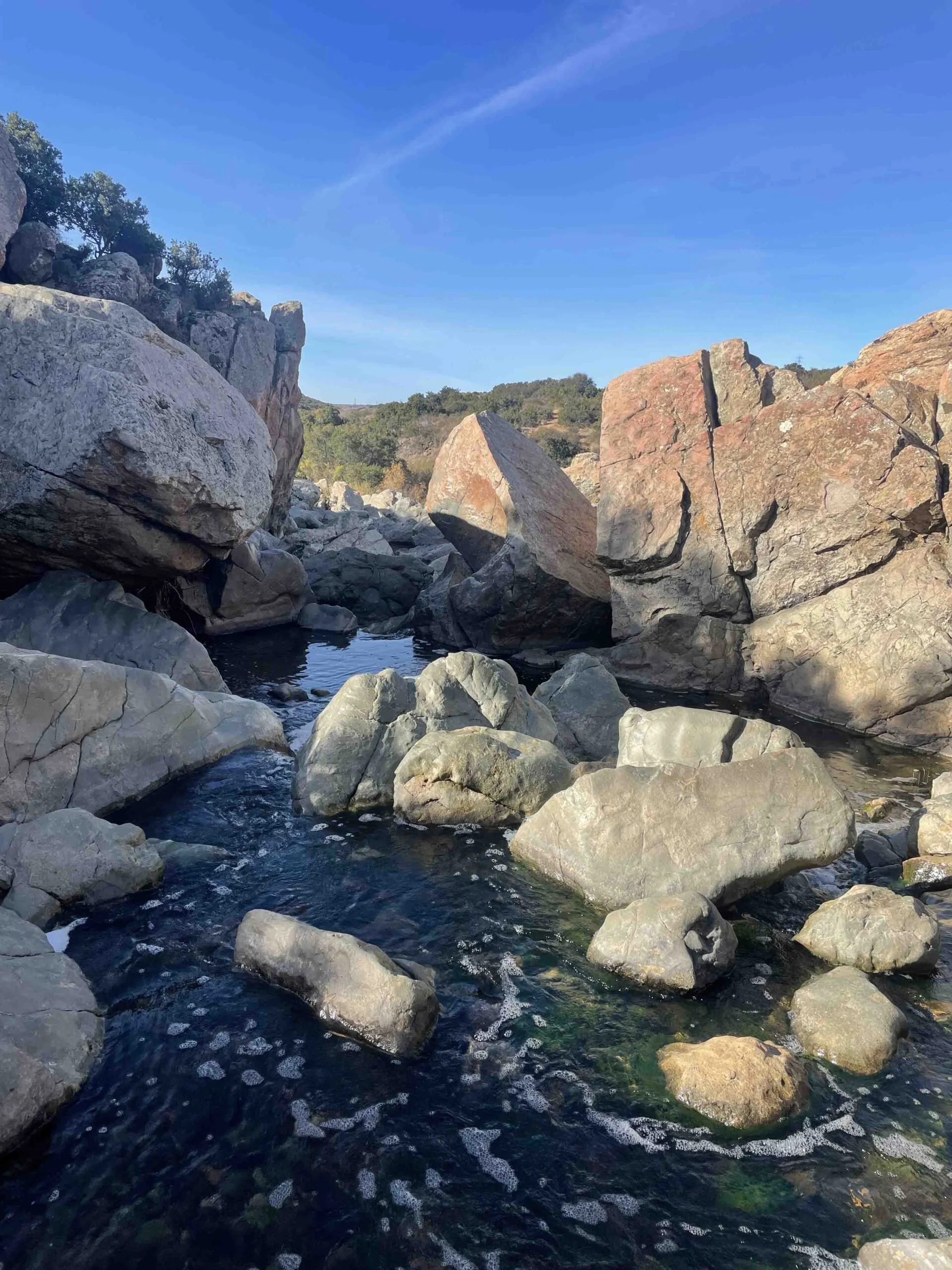 A rocky stream with large boulders and flowing water under a clear blue sky, with trees and hills in the background.