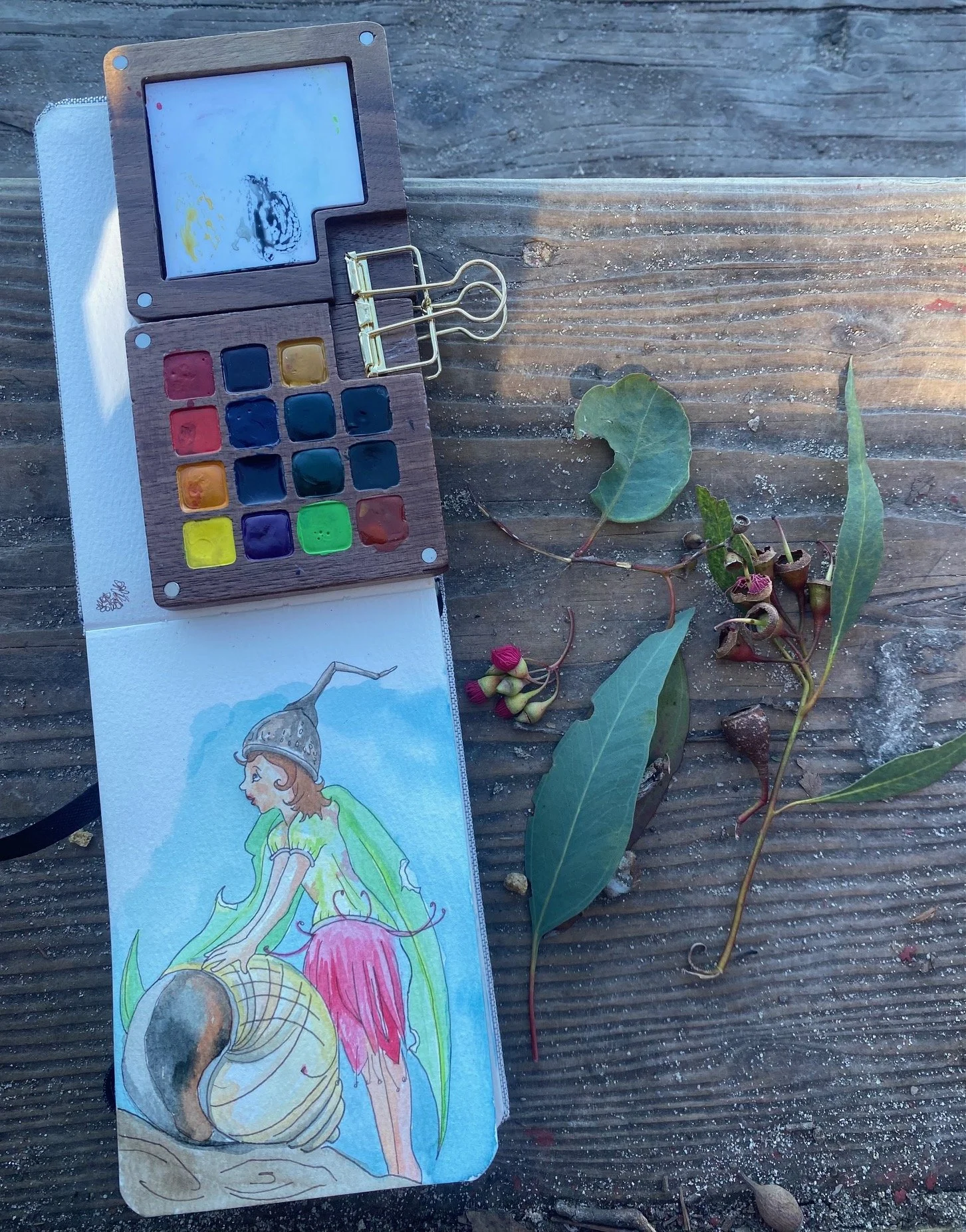 A watercolor painting of a fantasy character with a horned helmet, green cape, and red skirt, resting on a rock next to a snail shell, surrounded by leaves and small plants, on a wooden surface alongside a watercolor palette, paper towel, and some fallen leaves.