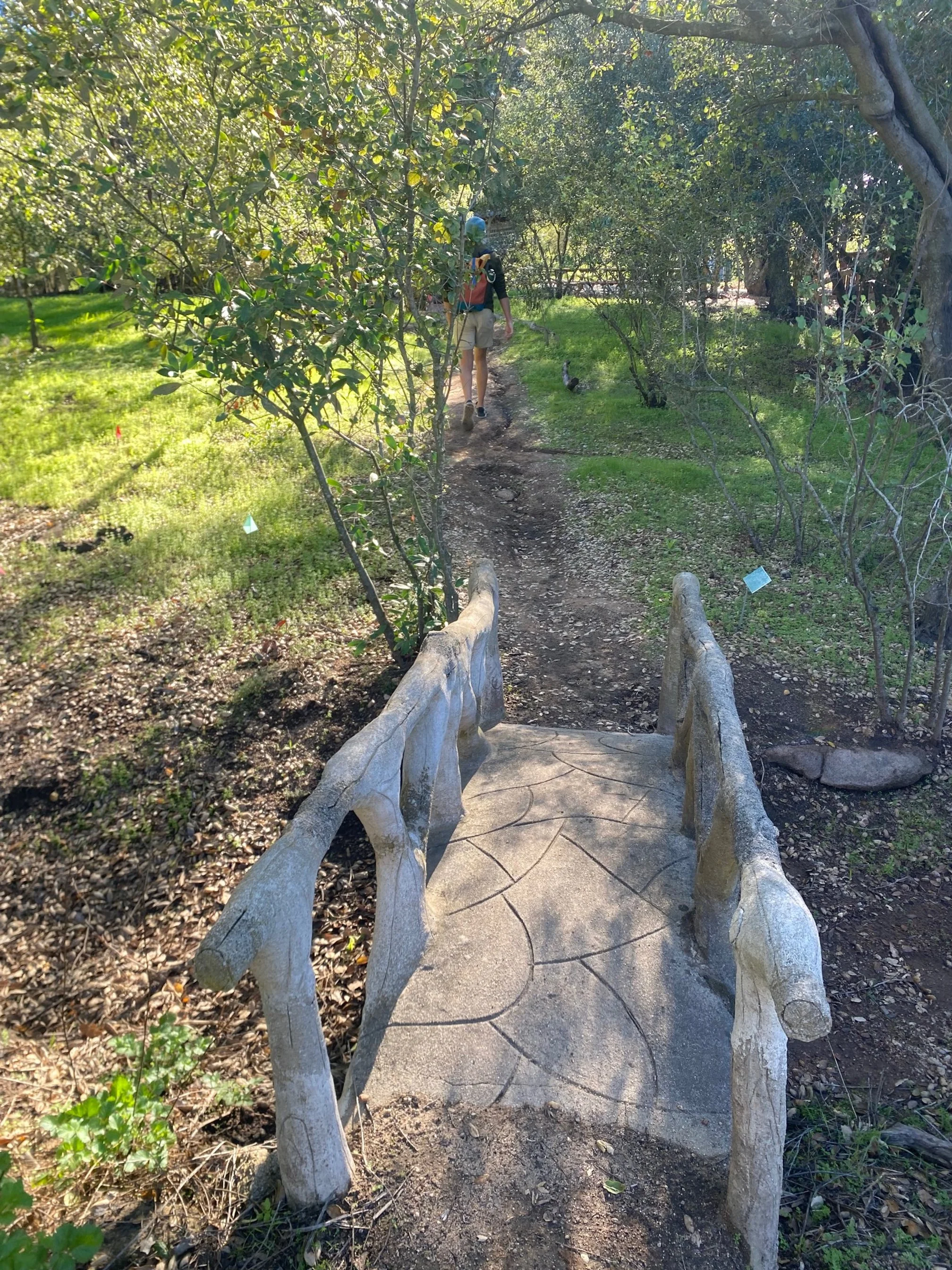 A small, decorative stone bridge with wooden railings spans a dirt path in a lush, green garden or park. A person is walking along the path, surrounded by trees and shrubbery.