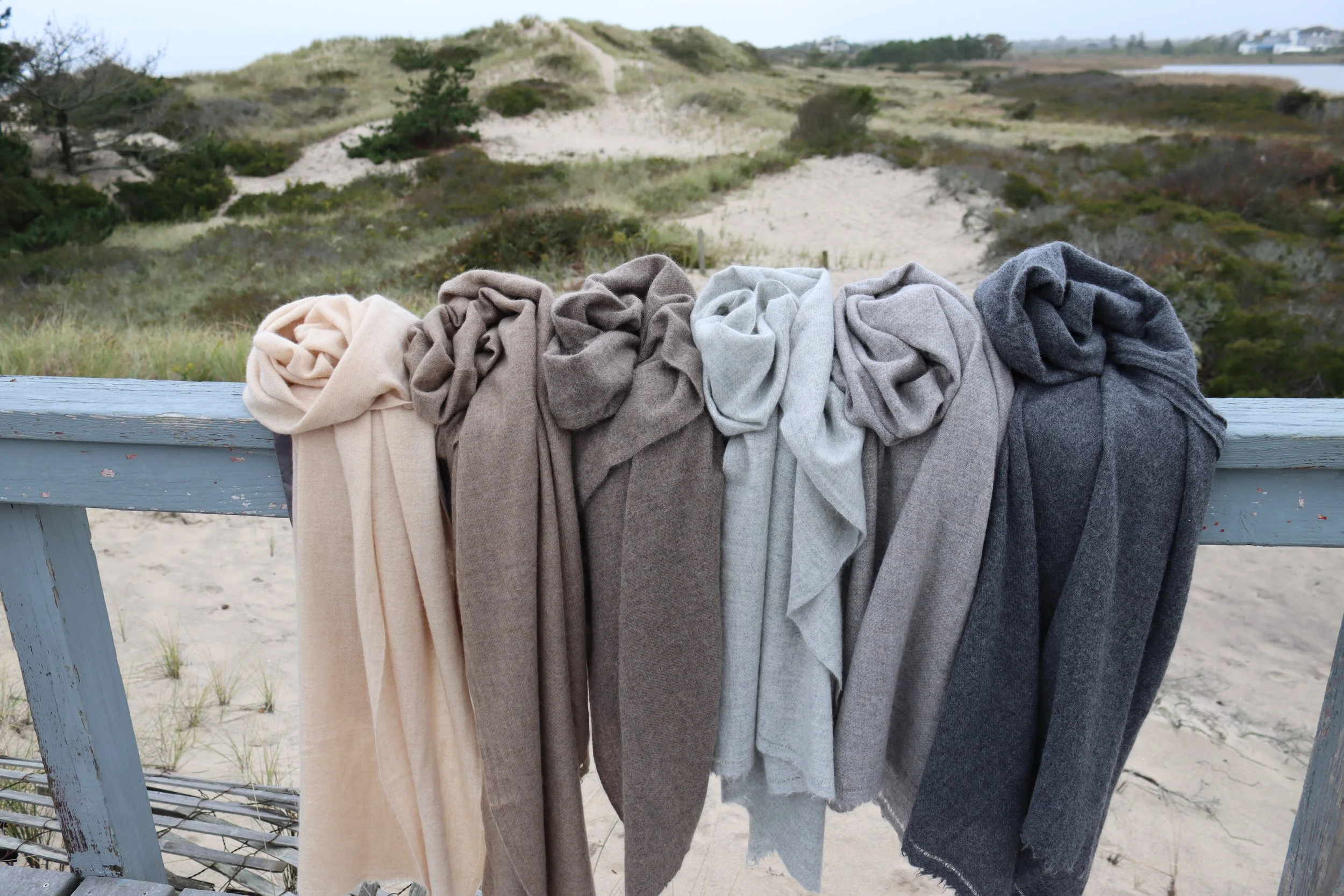 Multiple beige, brown, gray, and black cashmere shawls hung on a wooden railing at a Hamptons beach with dunes and sparse vegetation in the background.