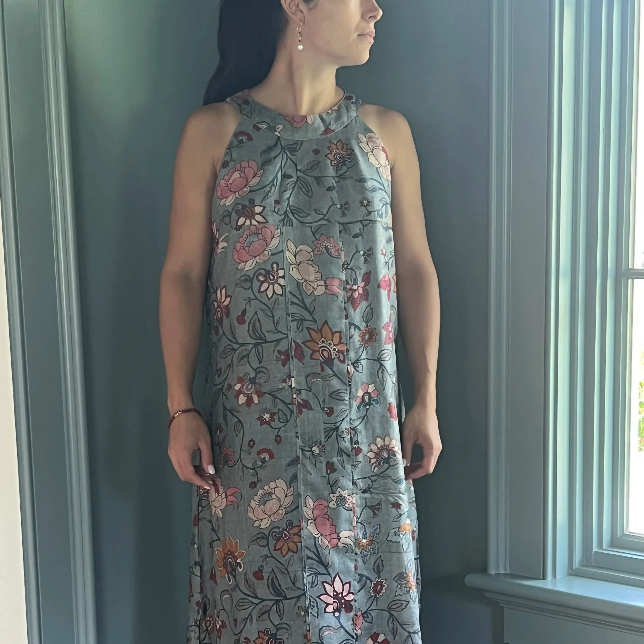 A woman in a floral dress stands near a window with green vertical blinds, facing to the side with her face partially visible.