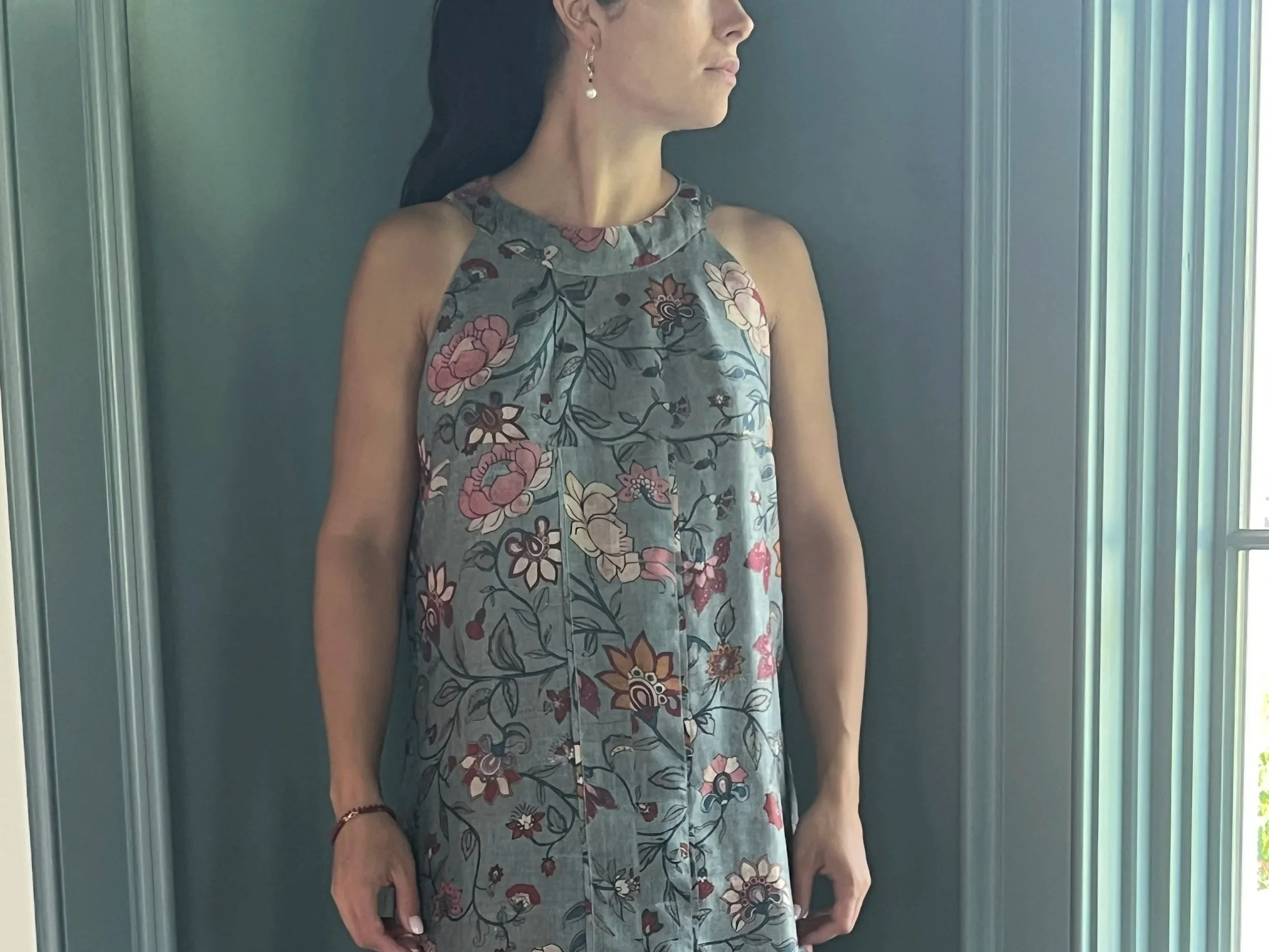 A woman wearing a sleeveless floral dress with a high neckline standing against a gray wall near a window with vertical blinds.