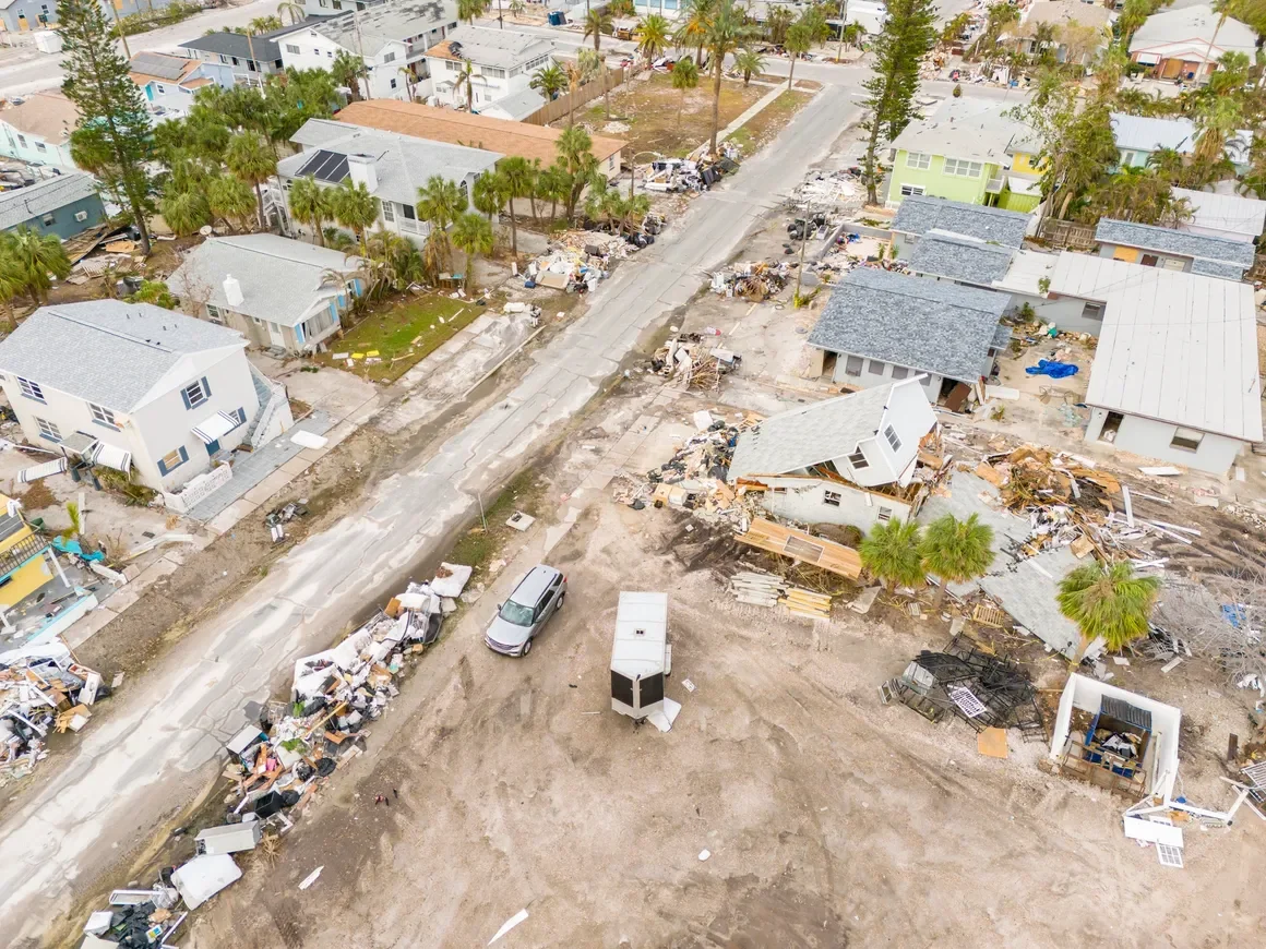 Aerial view of a neighborhood with houses damaged by a storm or hurricane, debris scattered on streets and yards, and some homes with roofs torn off.