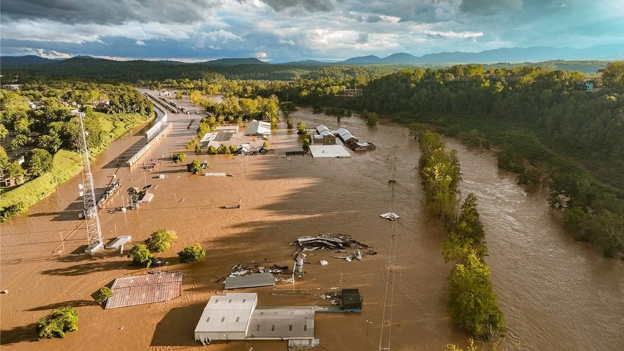 Aerial view of flooding with submerged buildings, trees, and roads, and damaged structures on muddy water, with hills and mountains in the background.