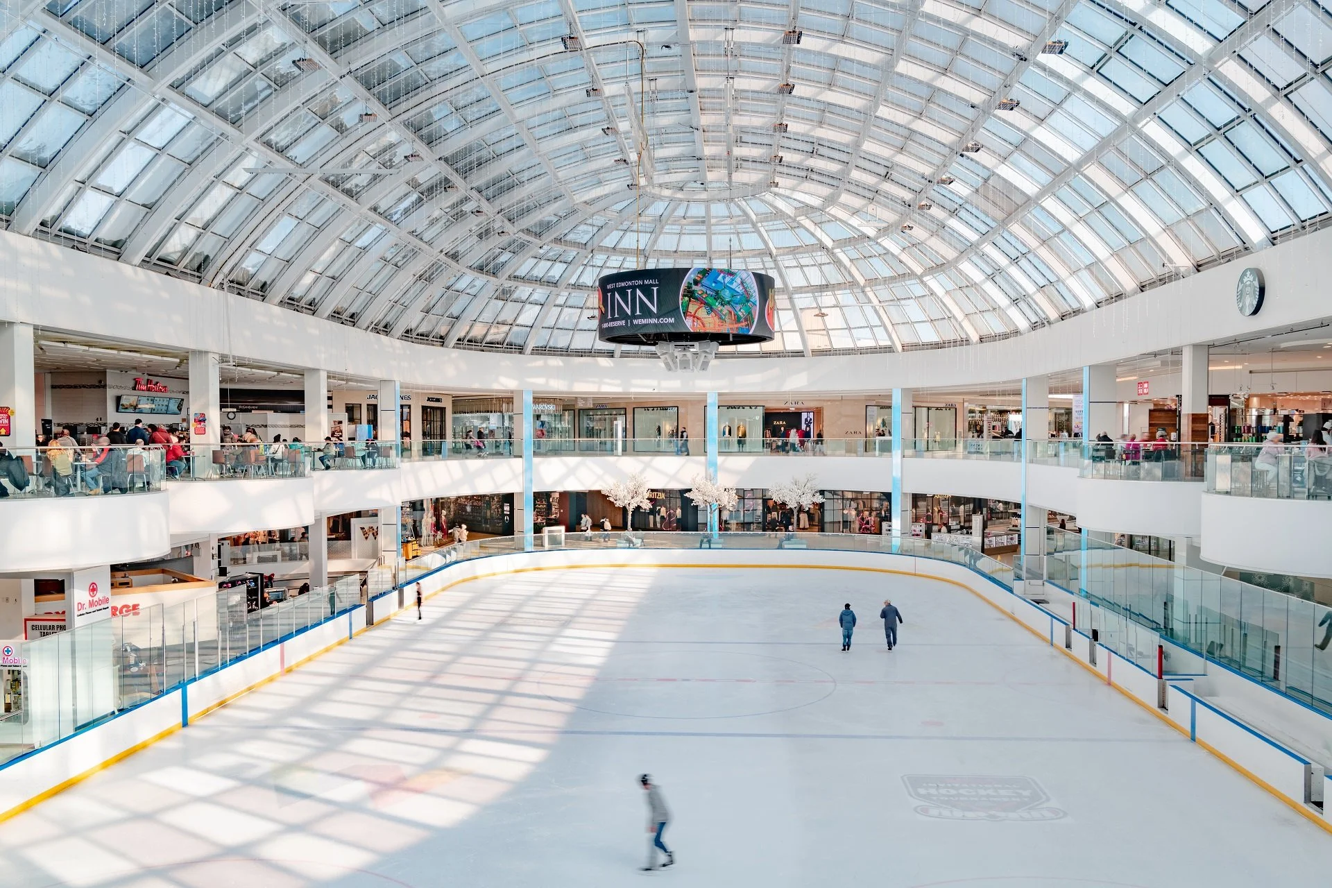 Indoor ice skating rink inside a shopping mall with a glass dome ceiling, surrounded by retail stores and seating areas, with a few skaters on the ice.