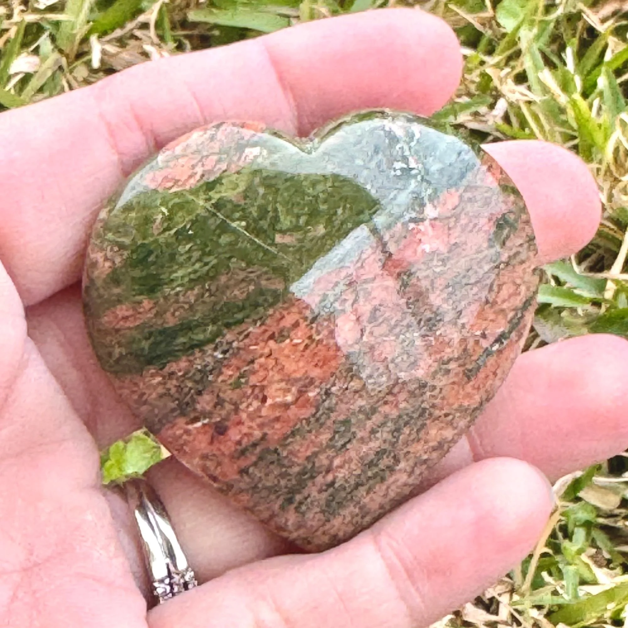 Hand holding a Unakite Jasper Heart featuring natural dark olive green and pink/red with grass in the background slightly blurred