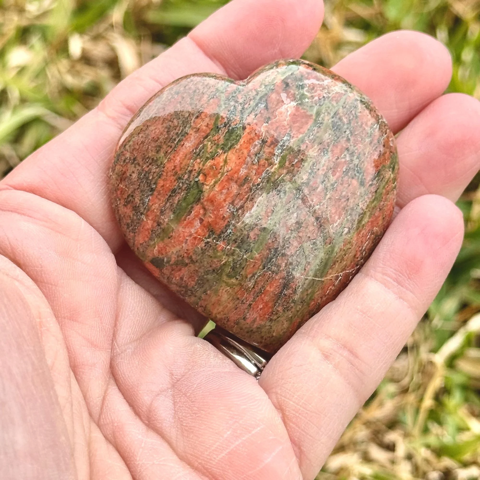 Hand holding a Unakite Jasper Heart featuring natural dark olive green and pink/red with grass in the background slightly blurred