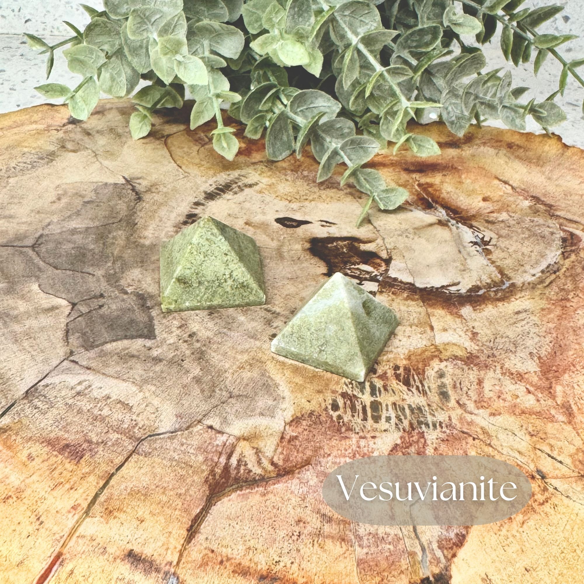 Two Vesuvianite Pyramid carvings in a olive green tone displayed on a petrified wood slab on a white countertop with a faux plant in the background