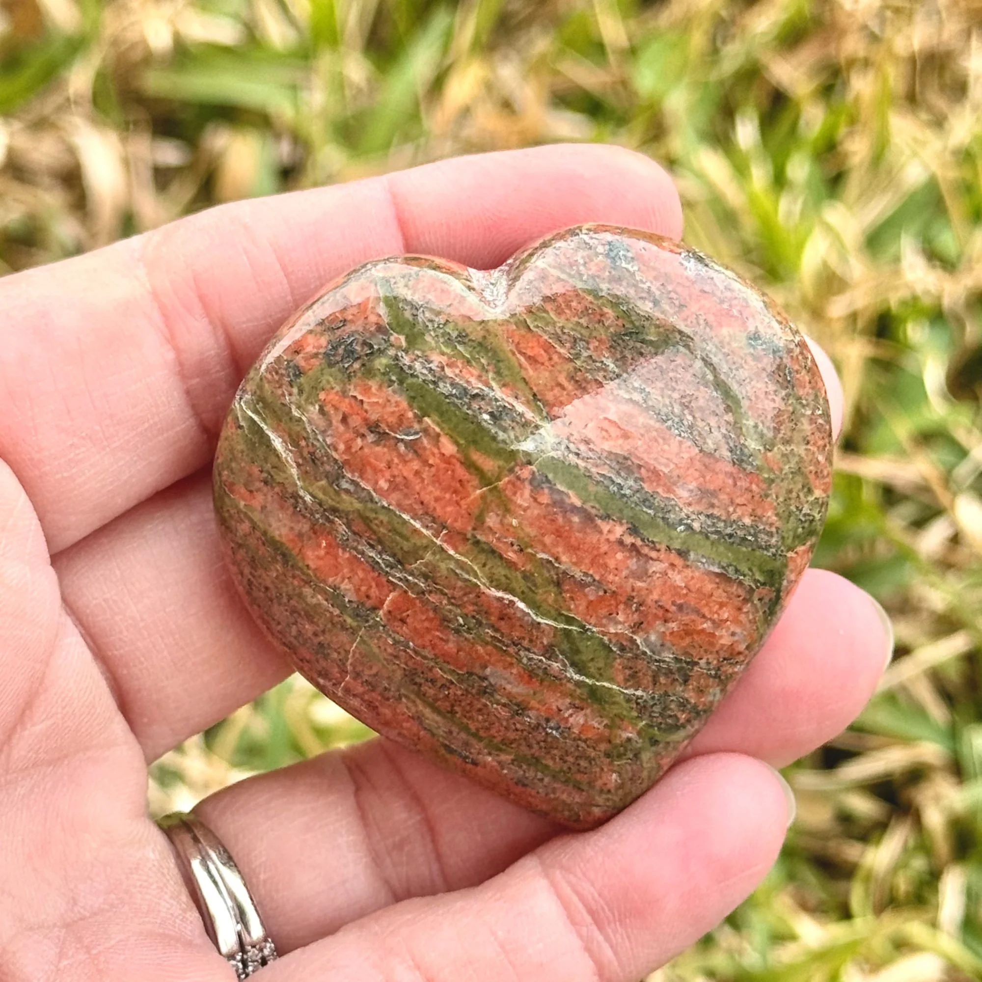 Hand holding a Unakite Jasper Heart featuring natural dark olive green and pink/red with grass in the background slightly blurred