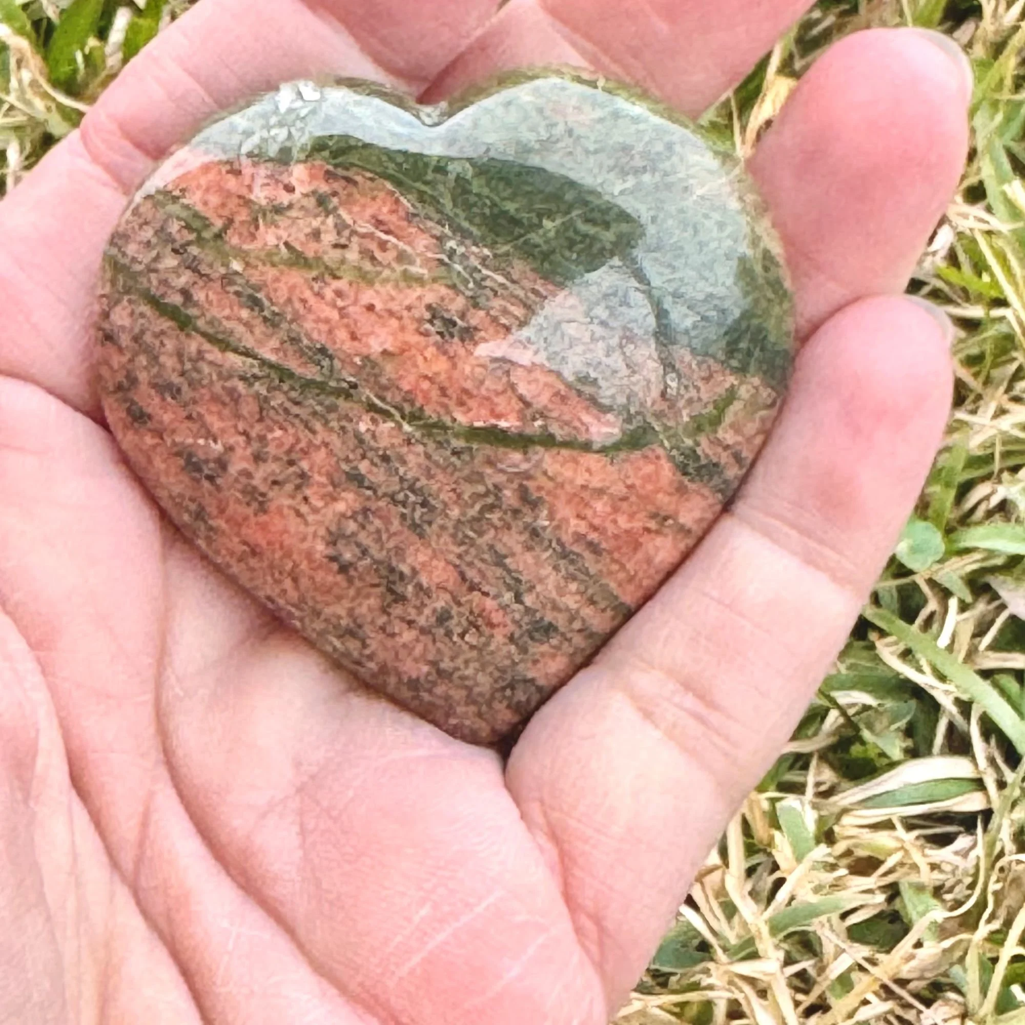 Hand holding a Unakite Jasper Heart featuring natural dark olive green and pink/red with grass in the background slightly blurred