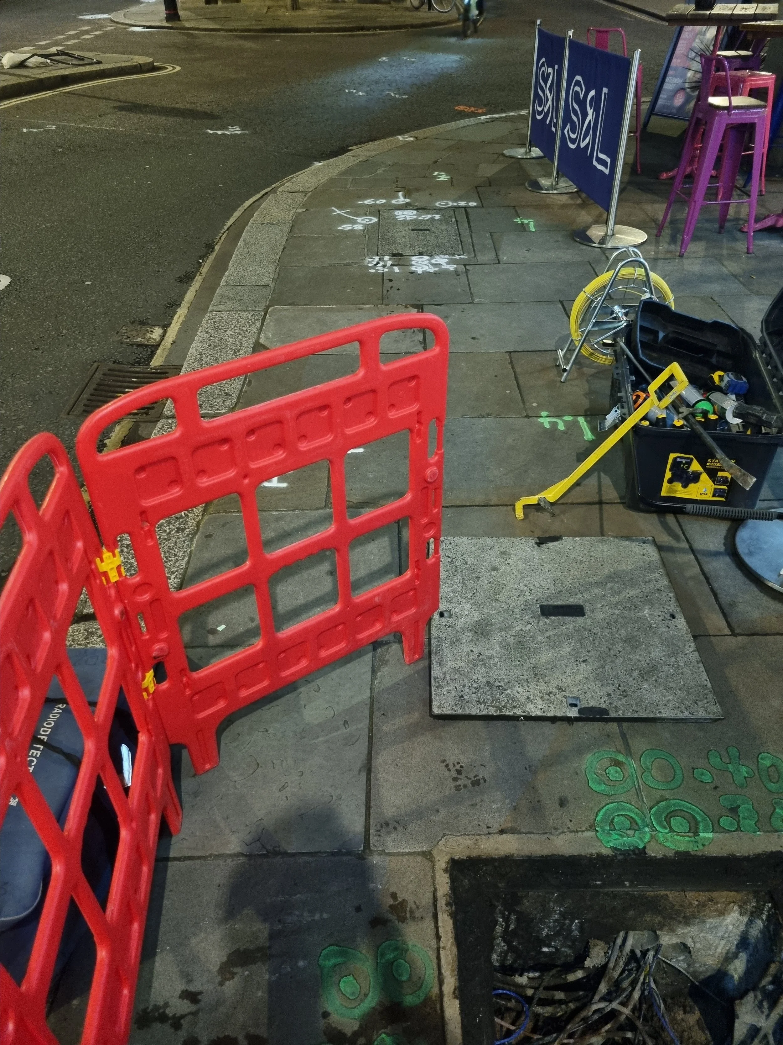 A sidewalk area under construction with traffic barriers, a toolbox, and marked utility access points on the ground.