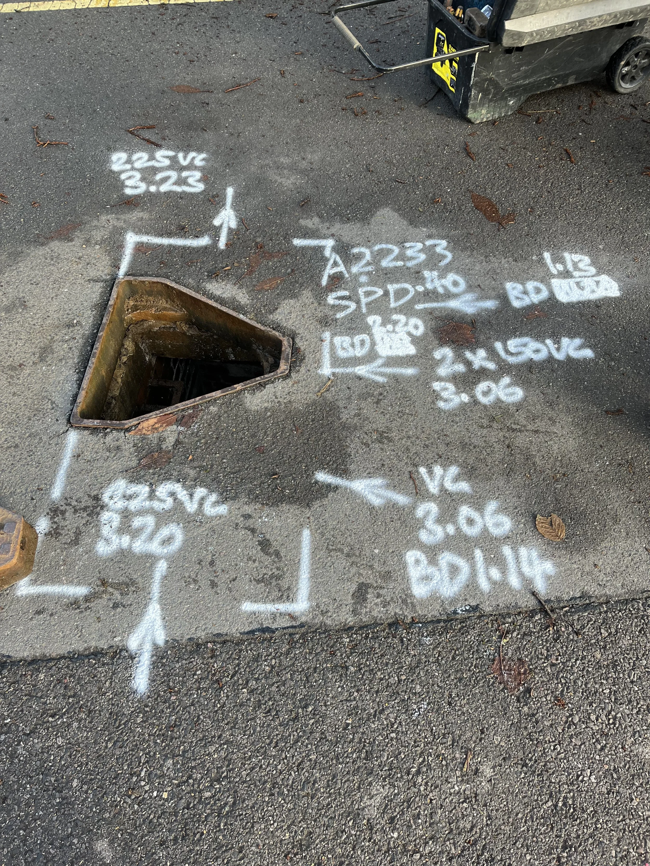 Hydro excavation site with white spray-painted markings indicating electrical and communication line locations and measurements around a manhole on asphalt.
