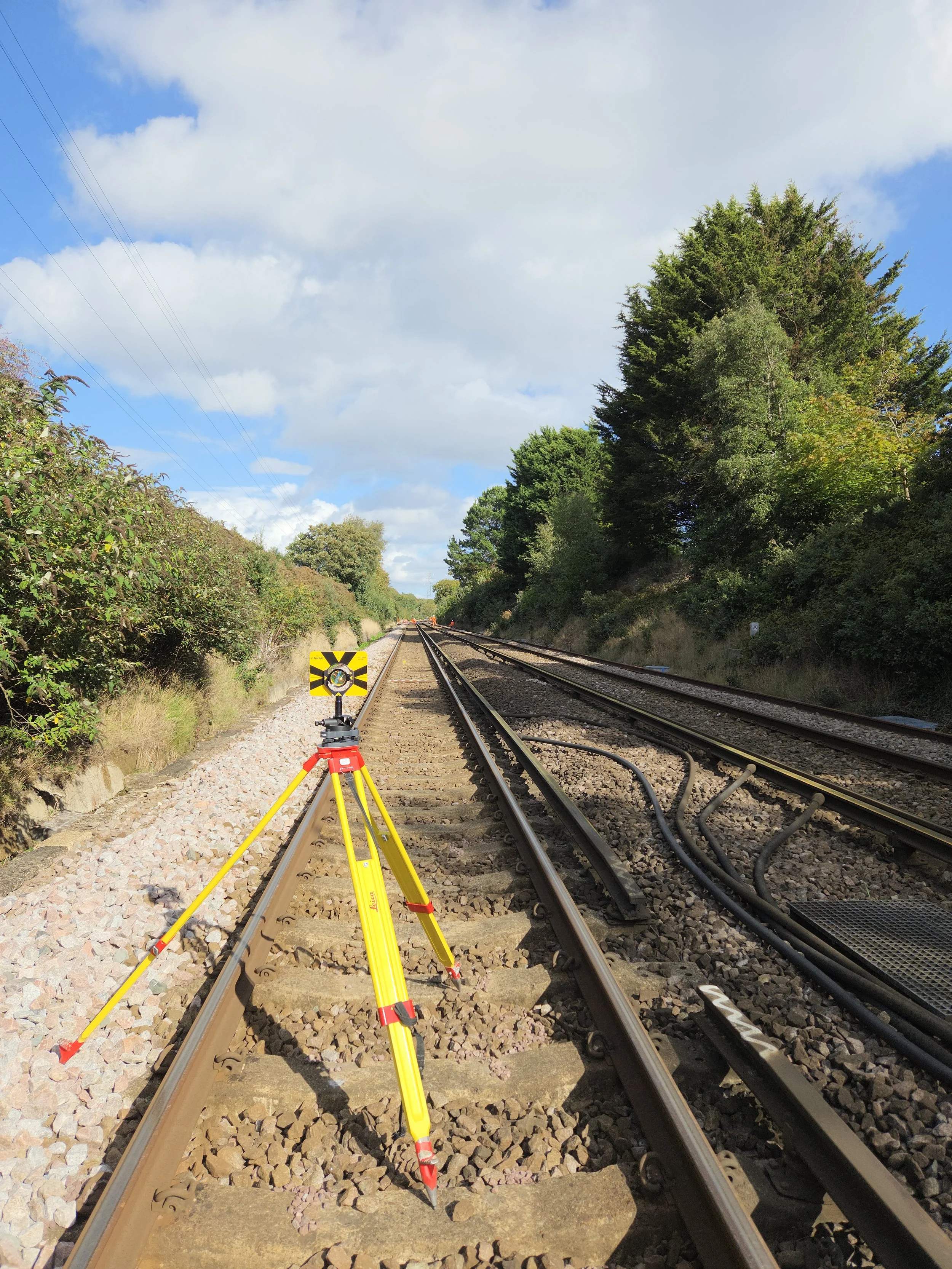 Surveying equipment set up on railroad tracks with trees on both sides and a partly cloudy sky.