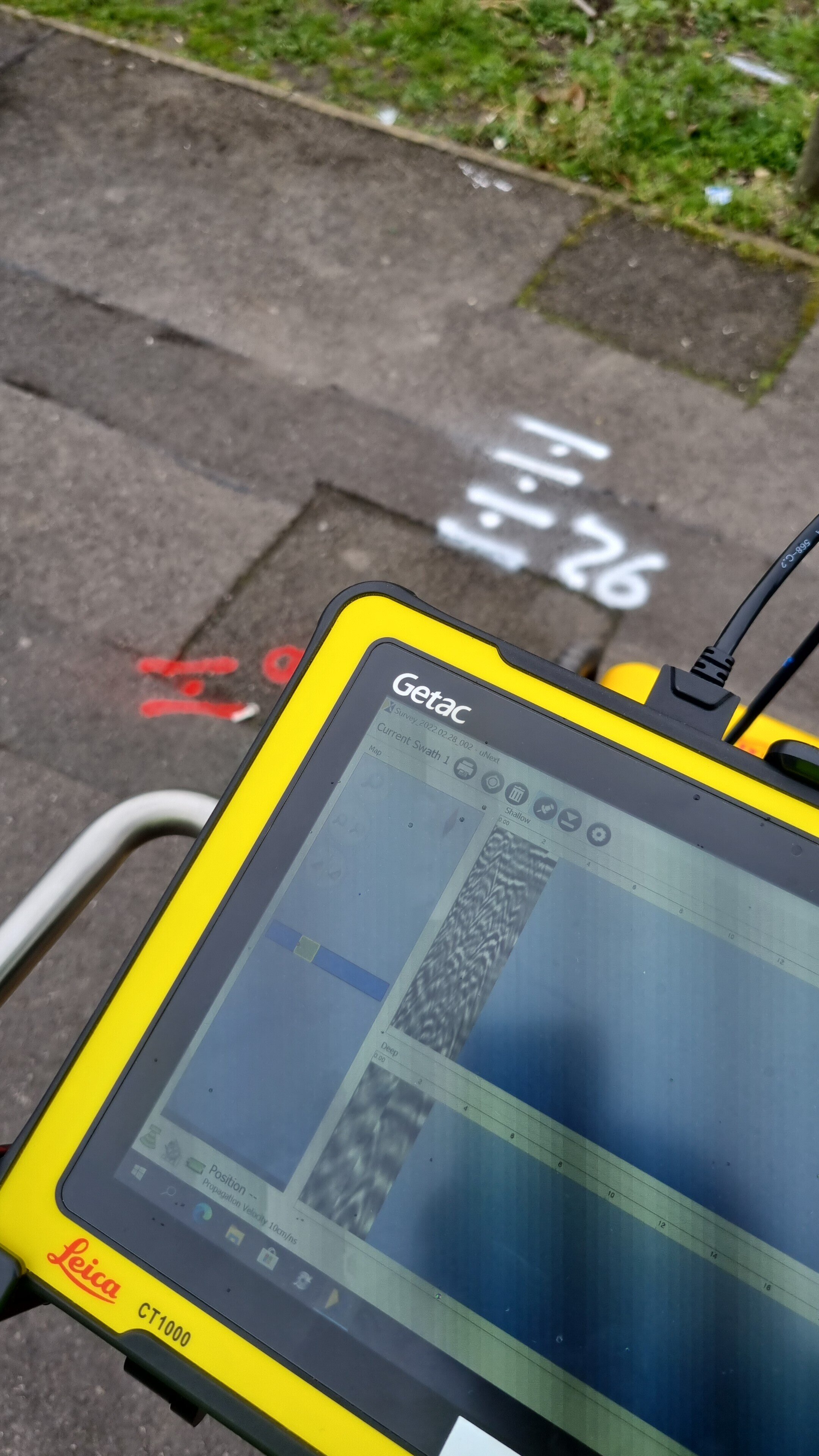Close-up of a yellow Leica surveying instrument displaying data on its screen, with a concrete sidewalk and green grass in the background, and white spray-painted markings on the pavement.