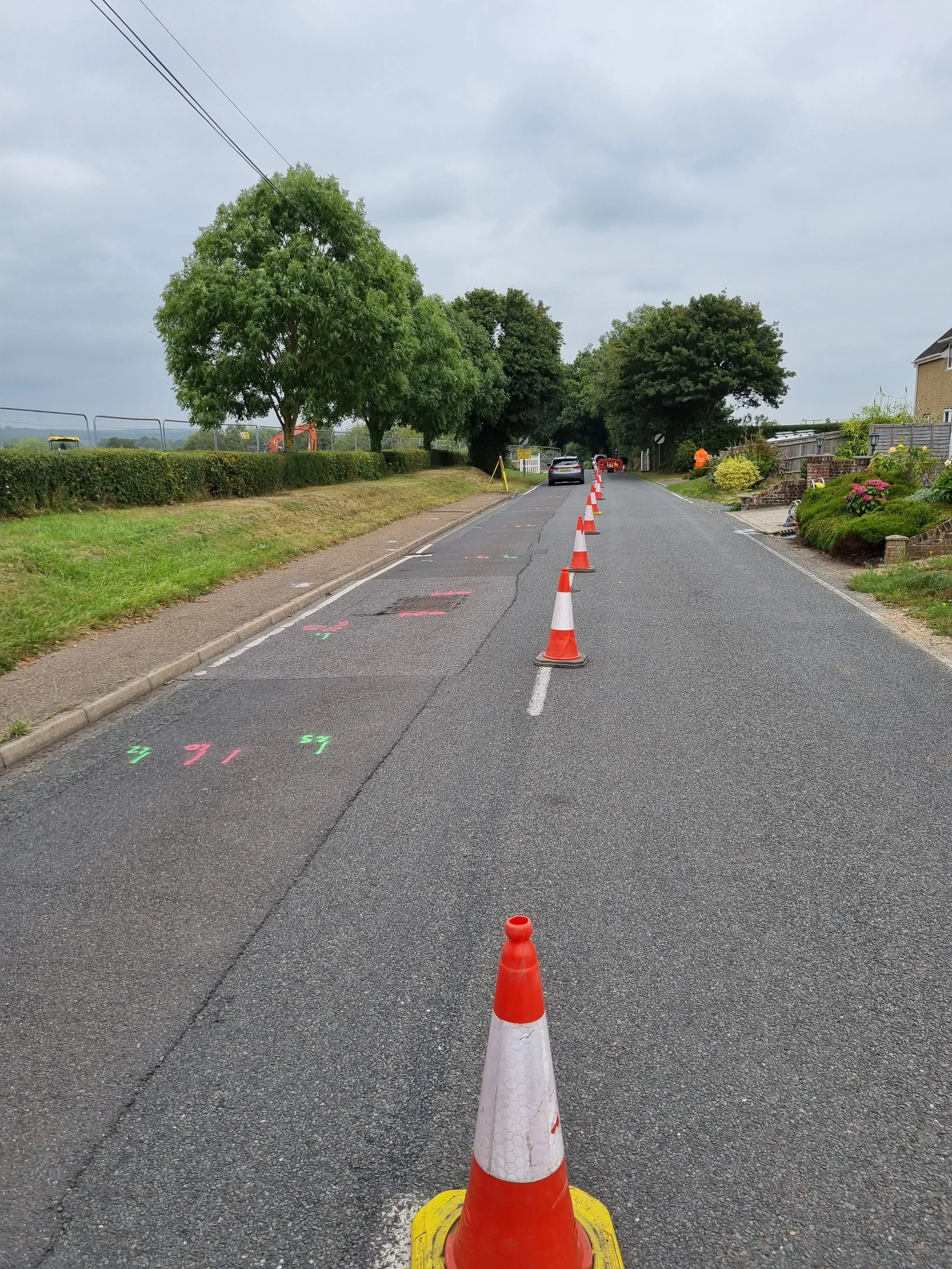 Roadwork or construction zone on a residential street with traffic cones and markings on the pavement. A car is parked further down the street, and construction equipment is visible in the background.