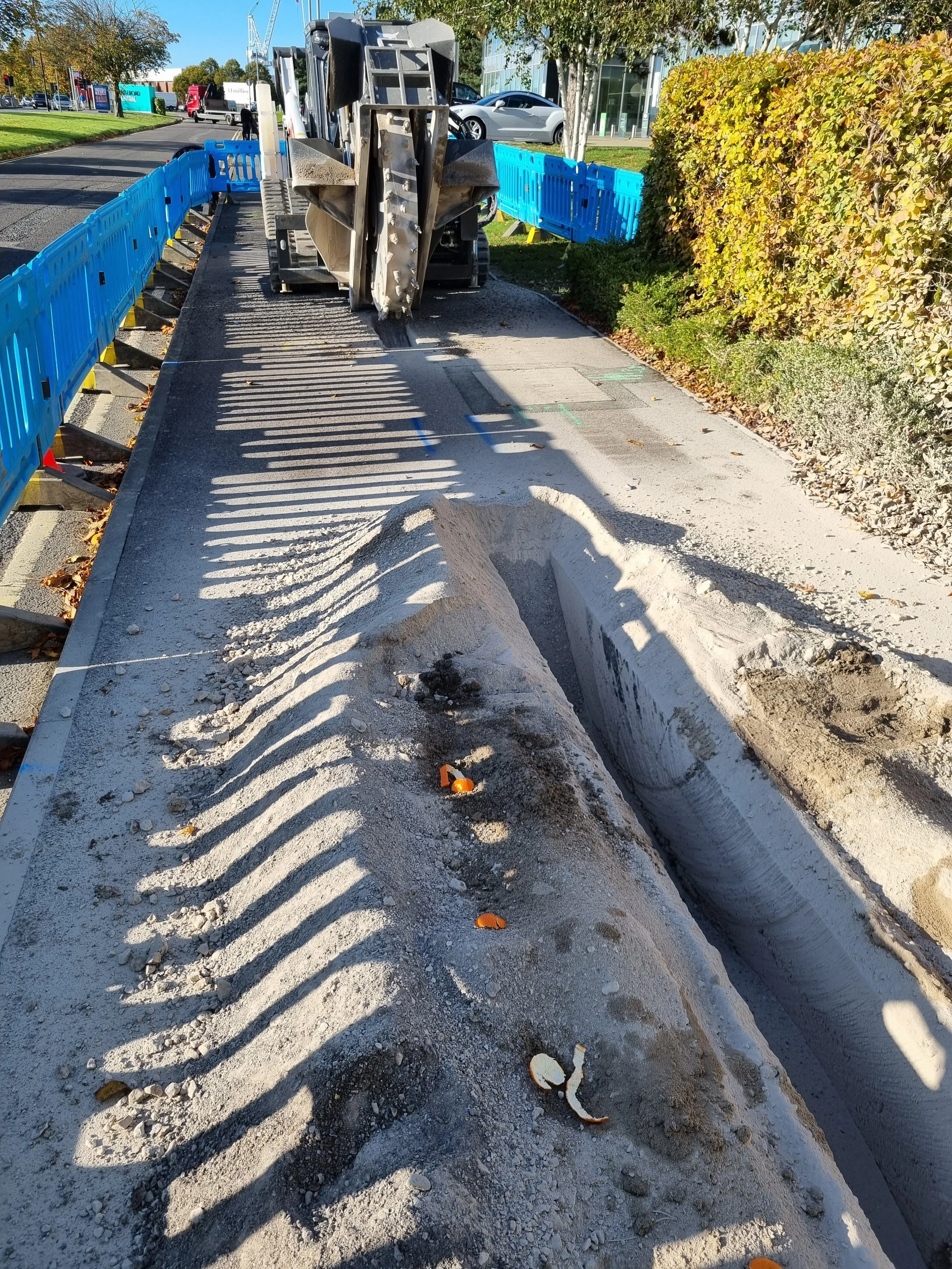 Construction site on a sidewalk with a large machine cutting into the pavement, surrounded by blue safety barriers and a hedge on the right side.