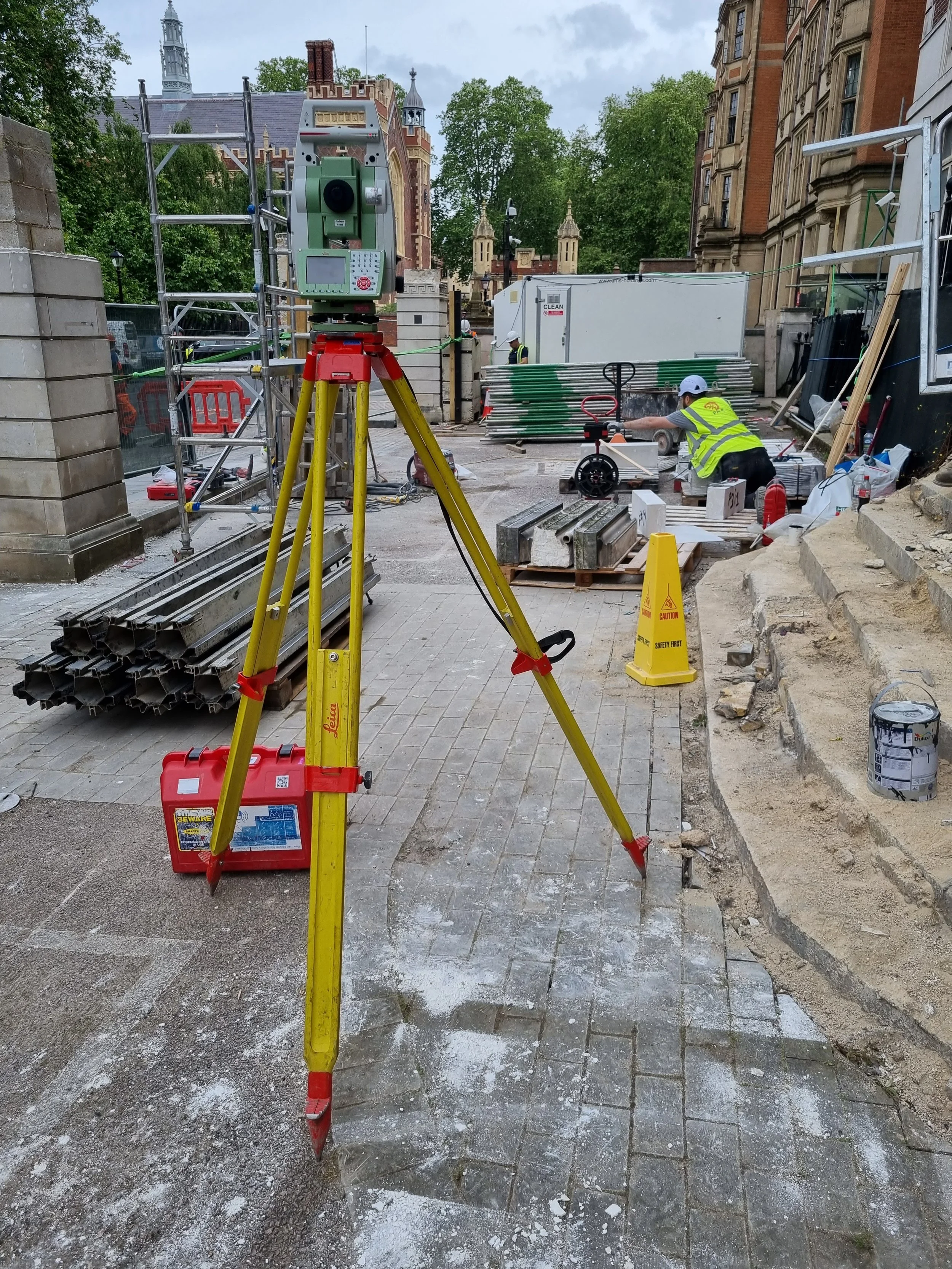 Construction site with a yellow surveyor tripod and equipment, workers in safety vests, construction materials, and safety signs, in front of historic buildings and a church with a clock tower.