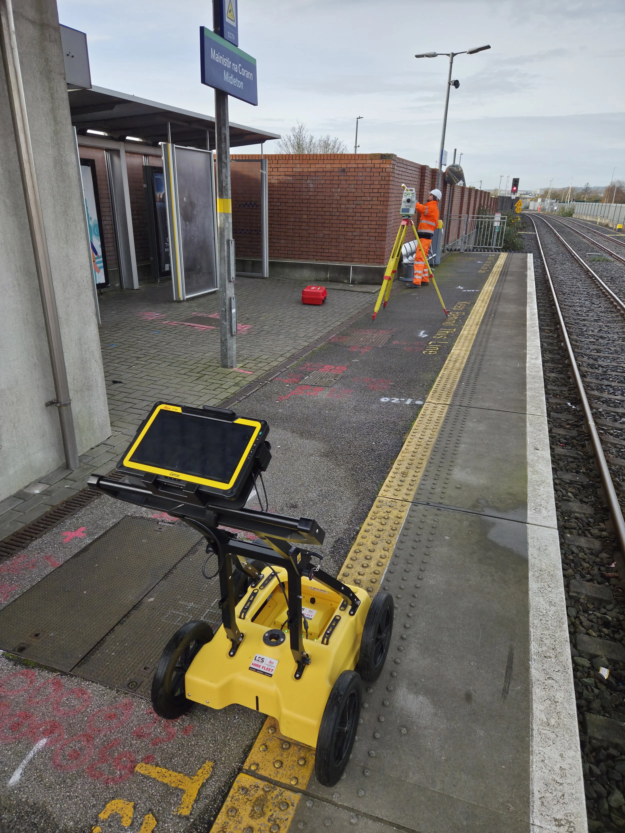 Railway platform with maintenance workers and surveying equipment, including a yellow robotic device in the foreground, a worker in orange safety gear operating a surveying instrument, and tracks extending into the distance.