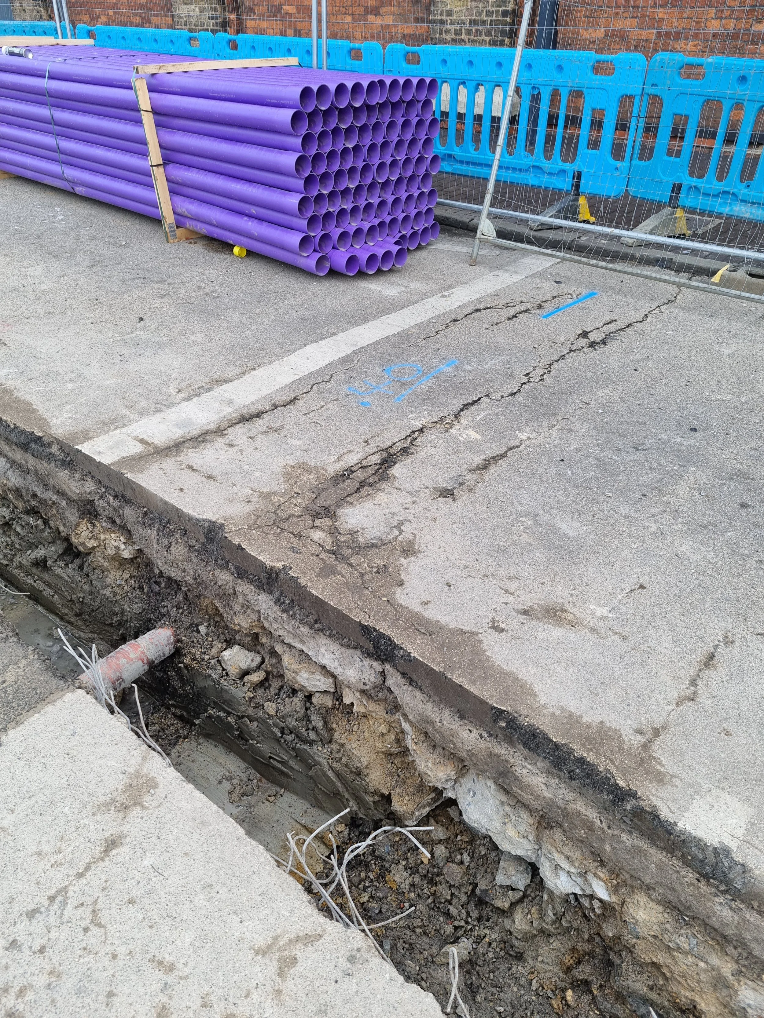Construction site with purple PVC pipes stacked on the sidewalk, blue safety barriers, and an open trench with exposed pipes and wiring.
