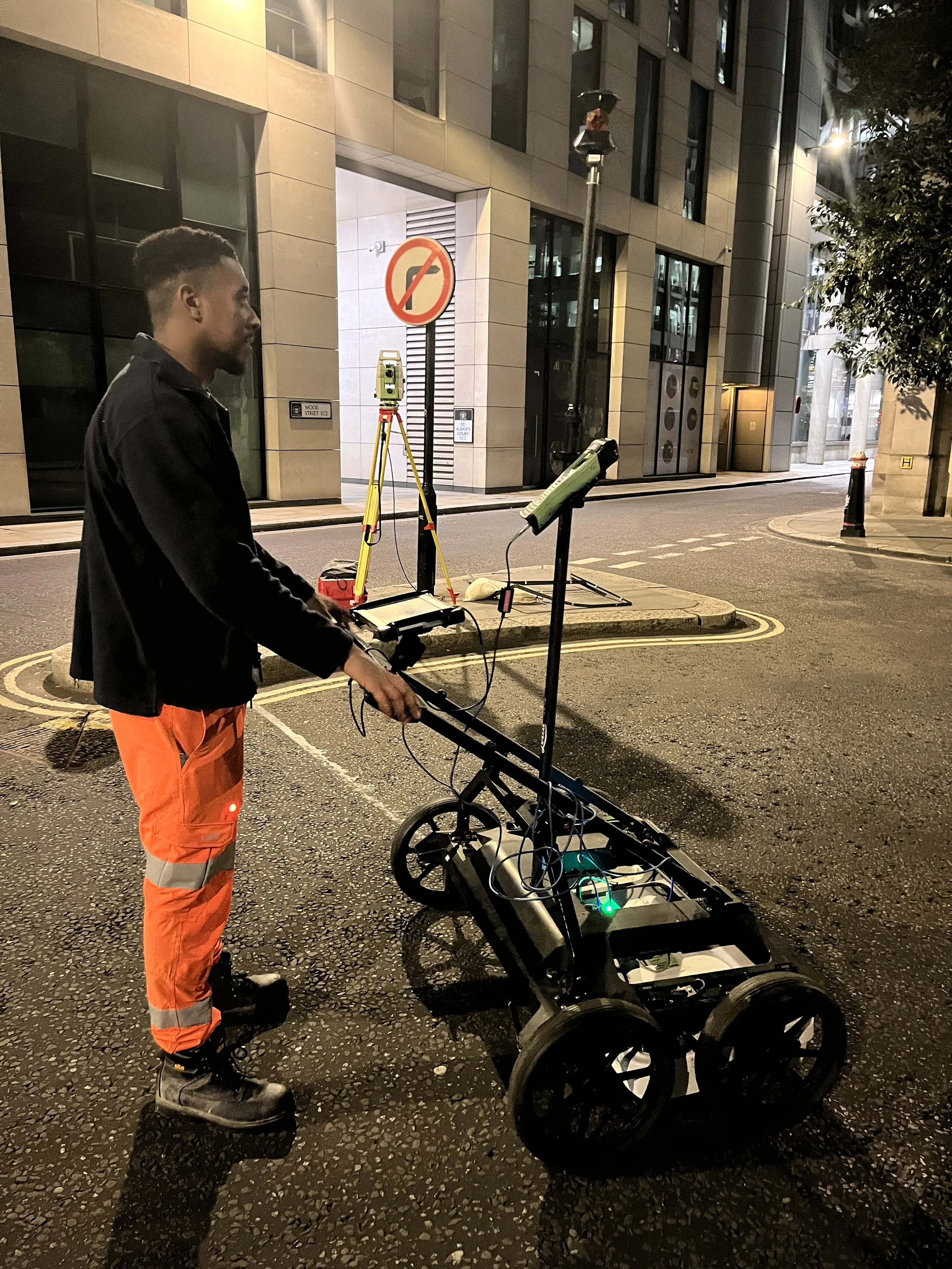 A man in orange work pants and black jacket operating a surveying instrument on a cart with wheels on a city street at night.