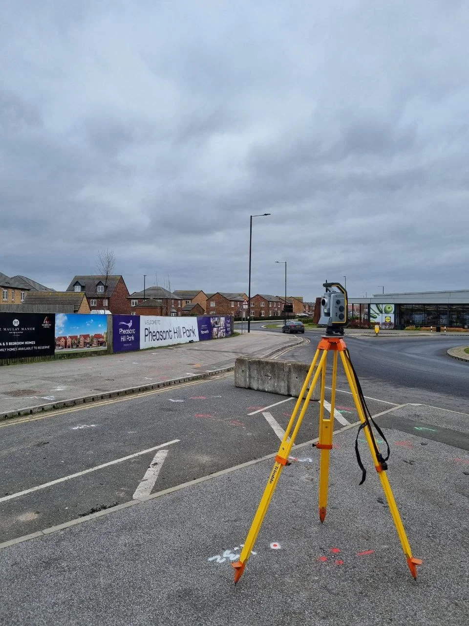 Surveying equipment set up on a parking lot, with residential neighborhood and cloudy sky in the background.