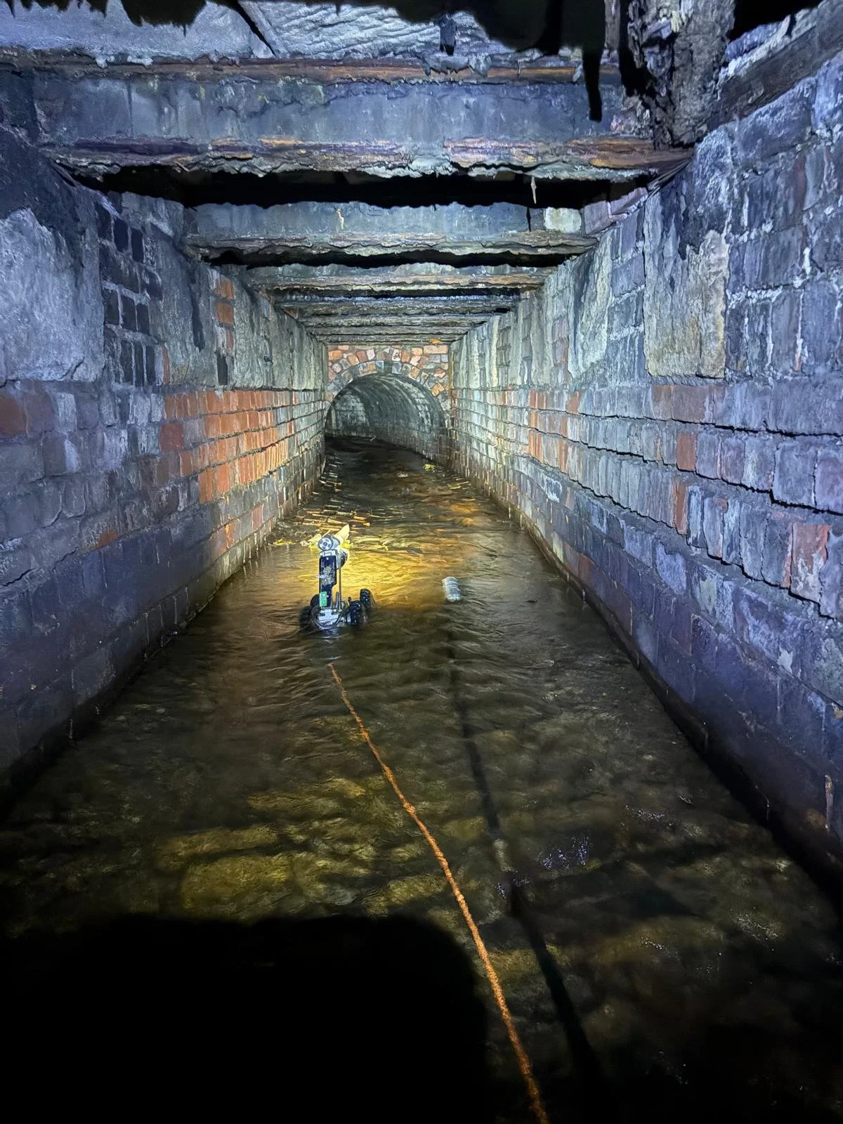 A submerged, narrow brick tunnel with water flowing through it, illuminated by a flashlight, with a small drone or robot floating in the water.