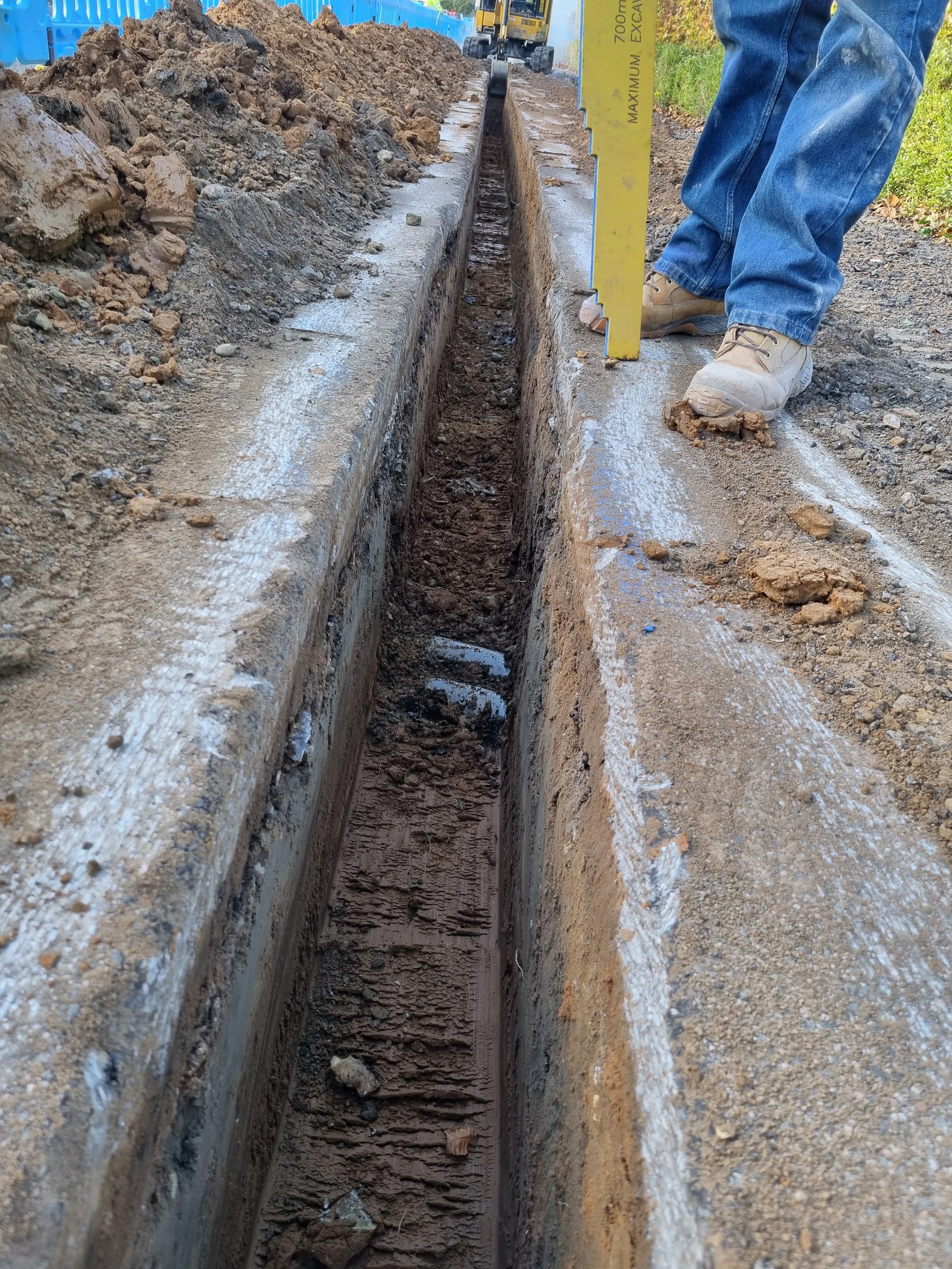 A construction worker is measuring the width of a freshly dug trench along a road with a yellow measuring stick. The worker is wearing jeans and tan work boots, and the trench contains some dirt and small debris.