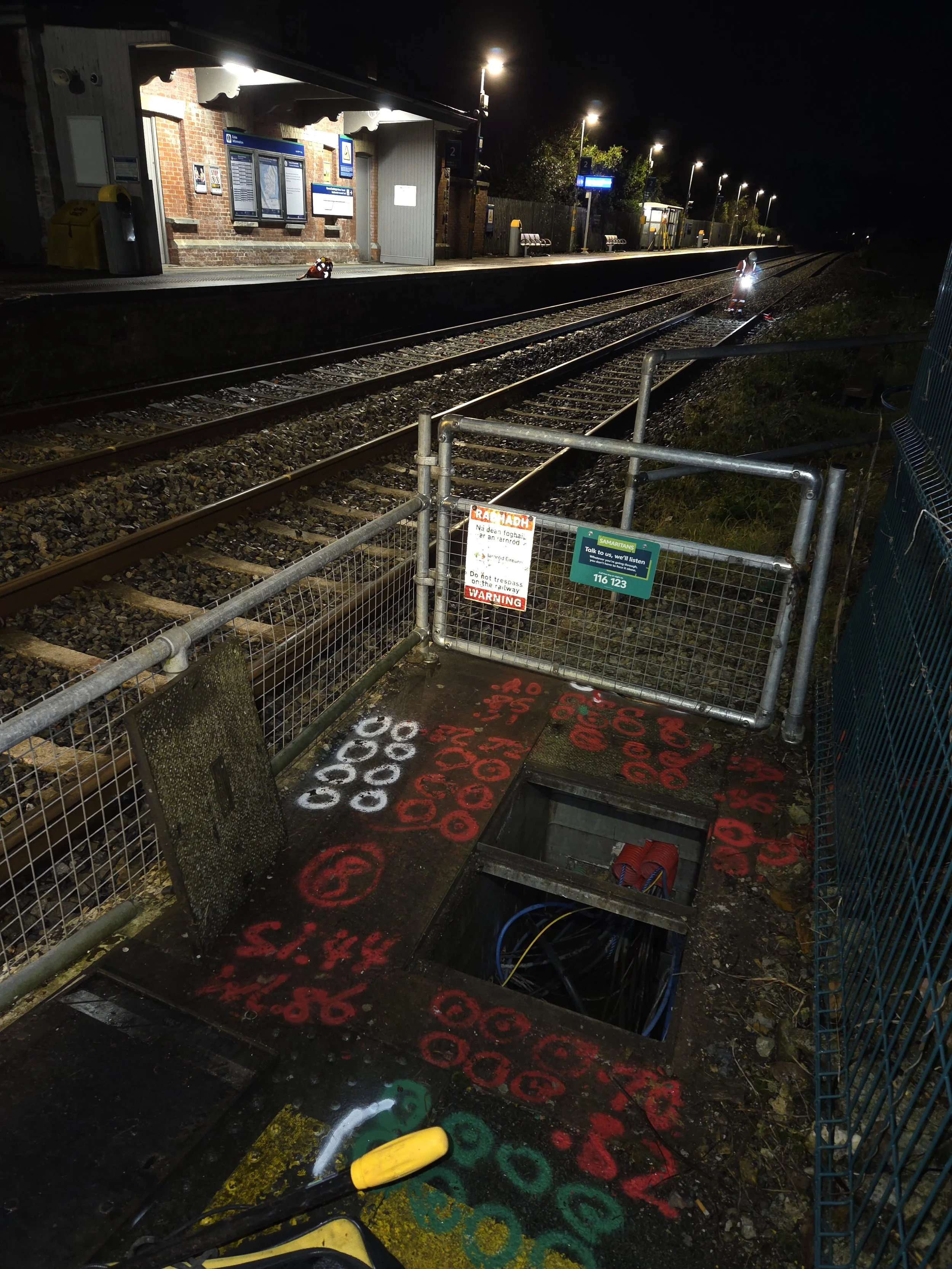Nighttime view of a train station platform with a closed gate in the foreground, graffiti on the ground, and train tracks extending into the distance illuminated by streetlights.