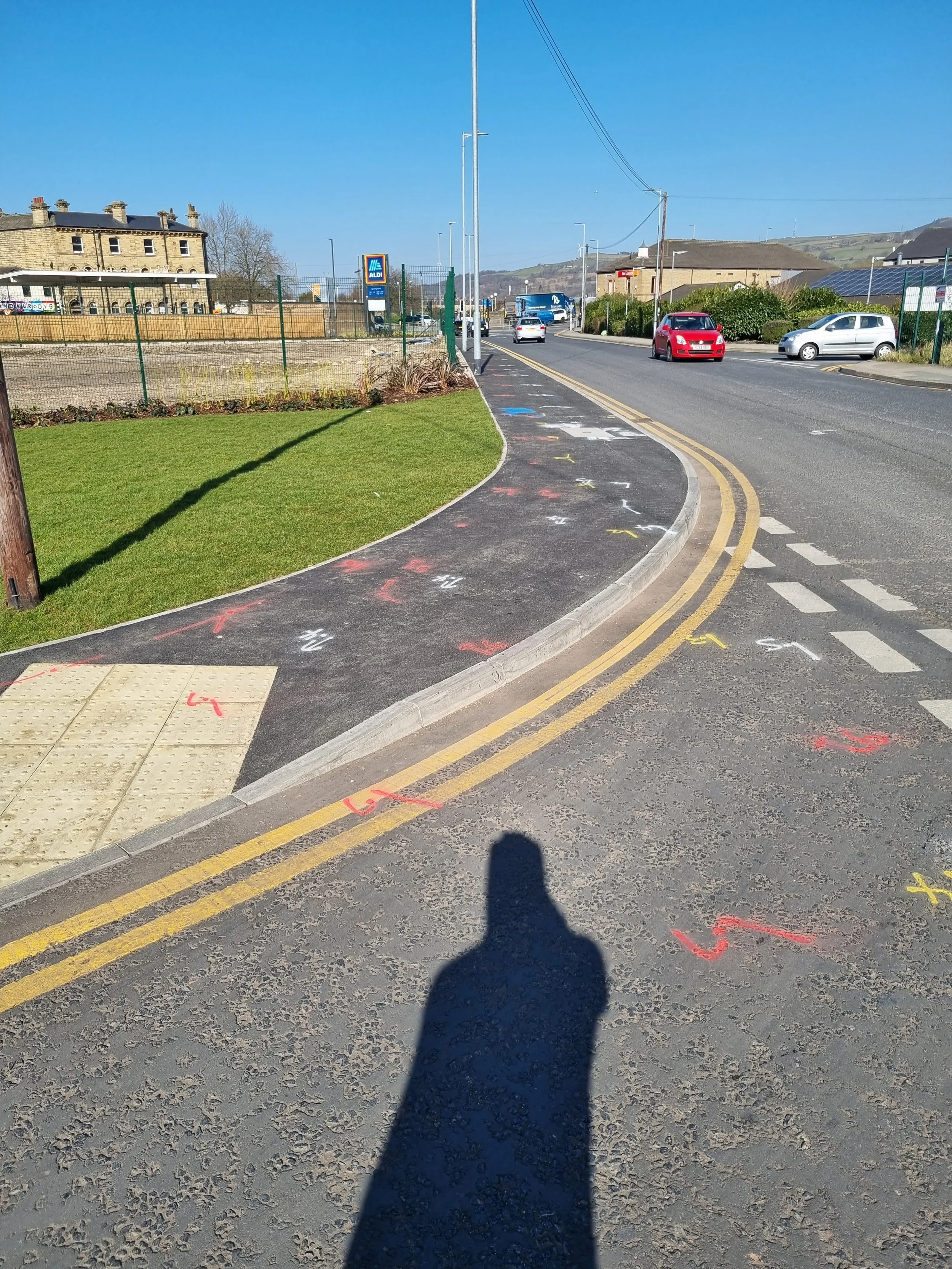 Sidewalk with colorful chalk markings, bordered by a curb and yellow double lines, with cars driving on a road and a grassy area on the side, under a clear blue sky.