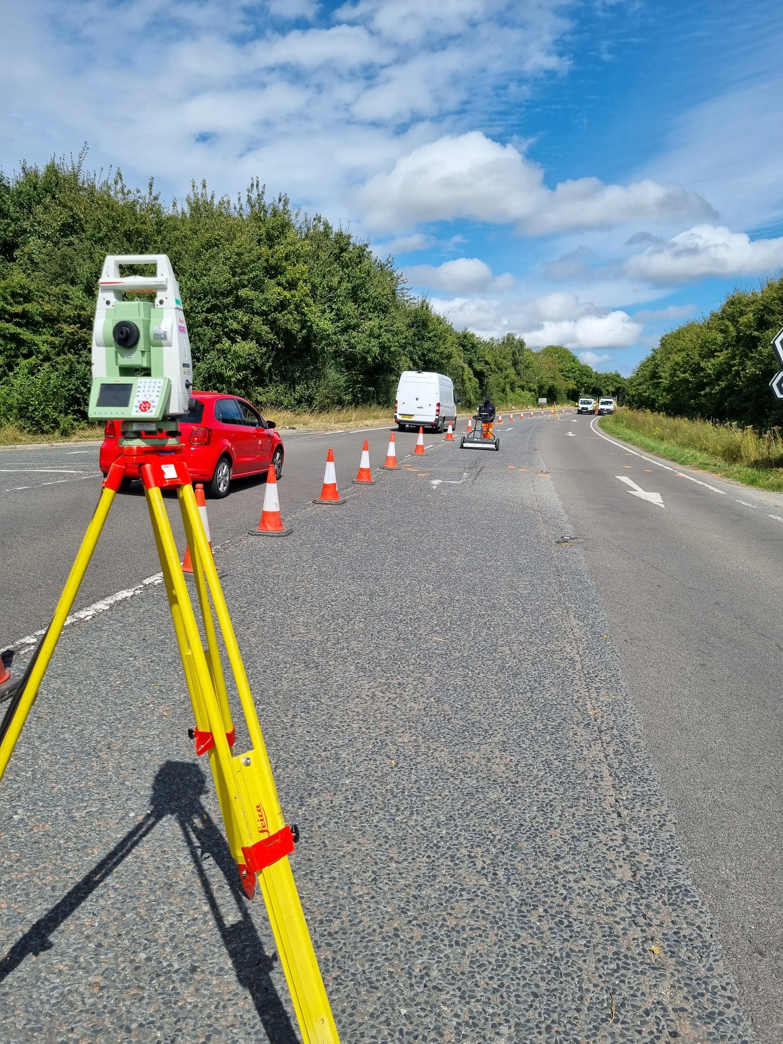 Road construction site with traffic cones, a surveying instrument on a tripod, and vehicles passing through a partially closed lane on a cloudy day.