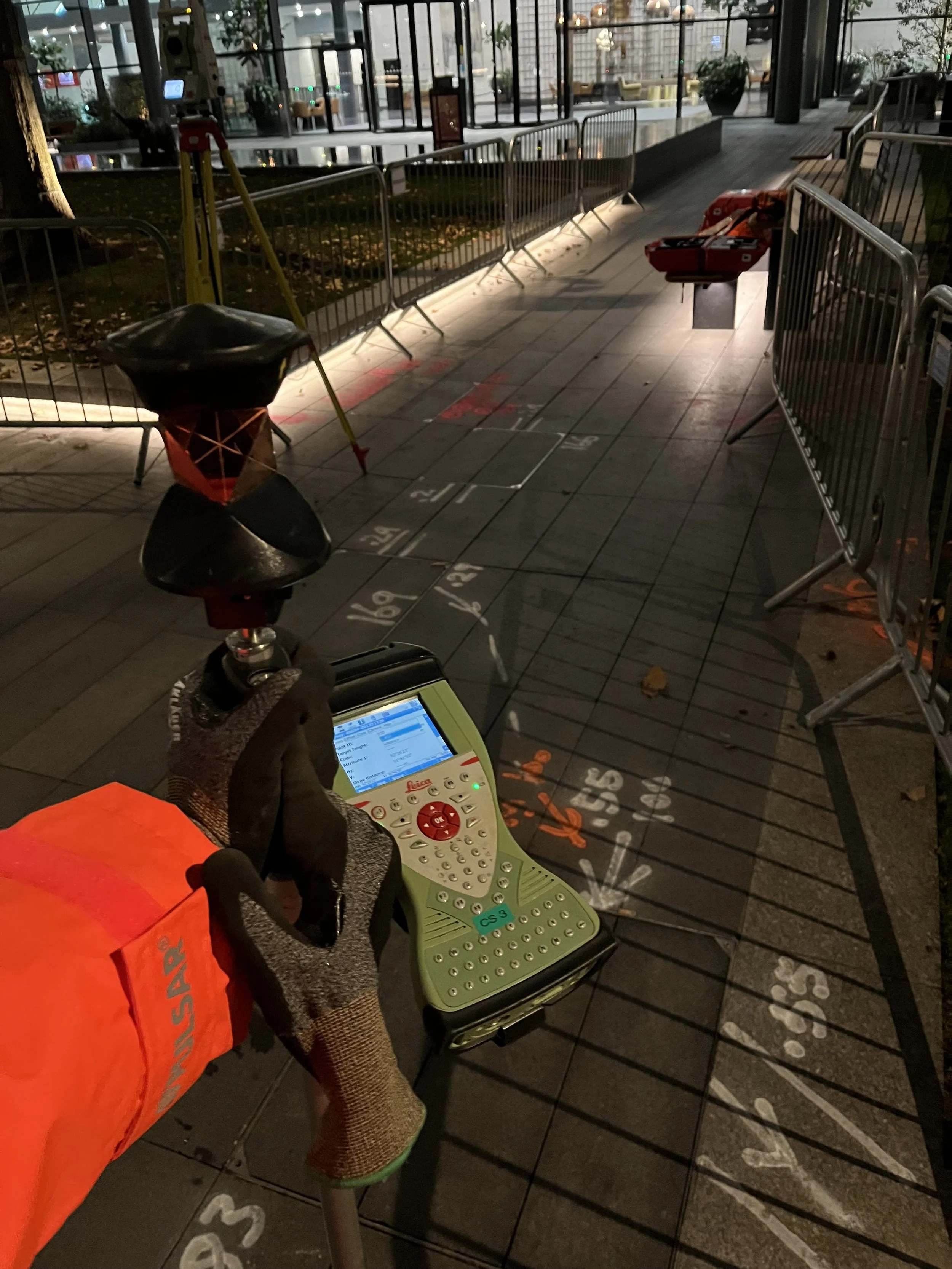 A person wearing safety gloves and a bright orange jacket holding a surveying instrument and a GPS device on a city sidewalk, with barriers and construction equipment visible in the background.